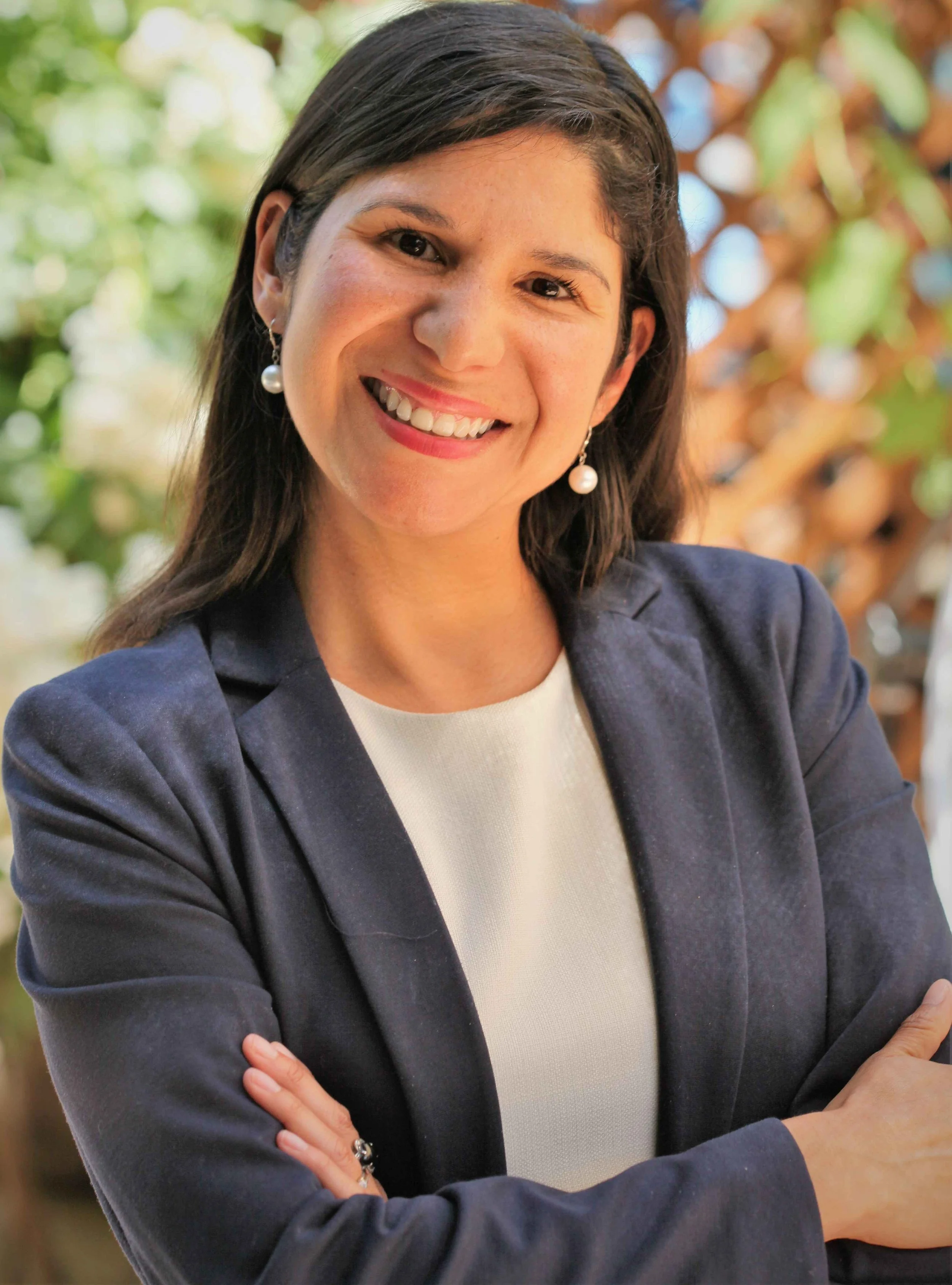 A woman with dark hair wearing pearl earrings, a navy blazer, and a white top, smiling with arms crossed, outdoors with blurred green and brown foliage in the background.