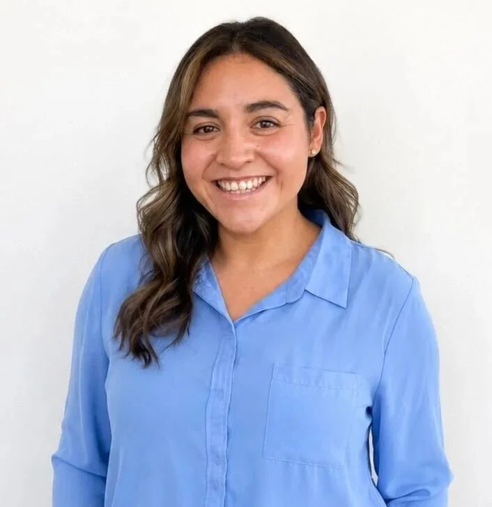 A woman with brown wavy hair smiling, wearing a light blue collared shirt, standing against a plain white background.