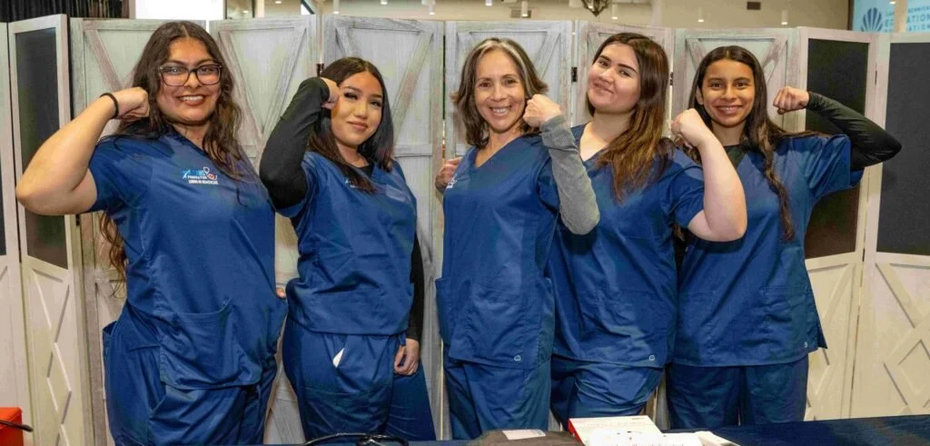 Five women in blue scrubs posing with flexed arms in front of a backdrop.