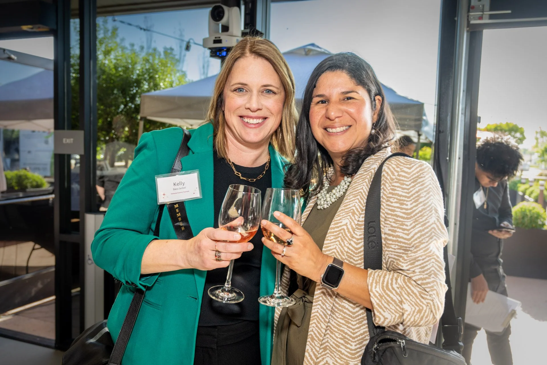 Two women smiling and holding glasses of wine at an outdoor event, dressed professionally, with tents and trees in the background.
