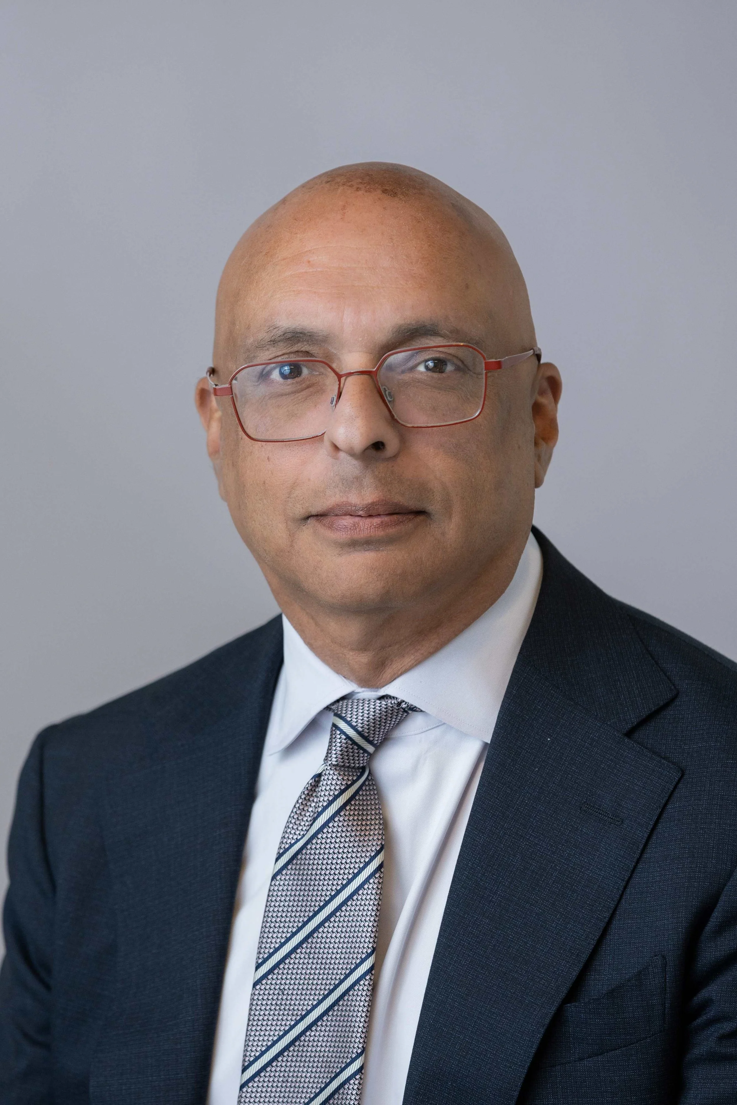 A professional headshot of a man with a shaved head and glasses, wearing a dark suit, white shirt, and patterned tie, standing against a plain, light gray background.