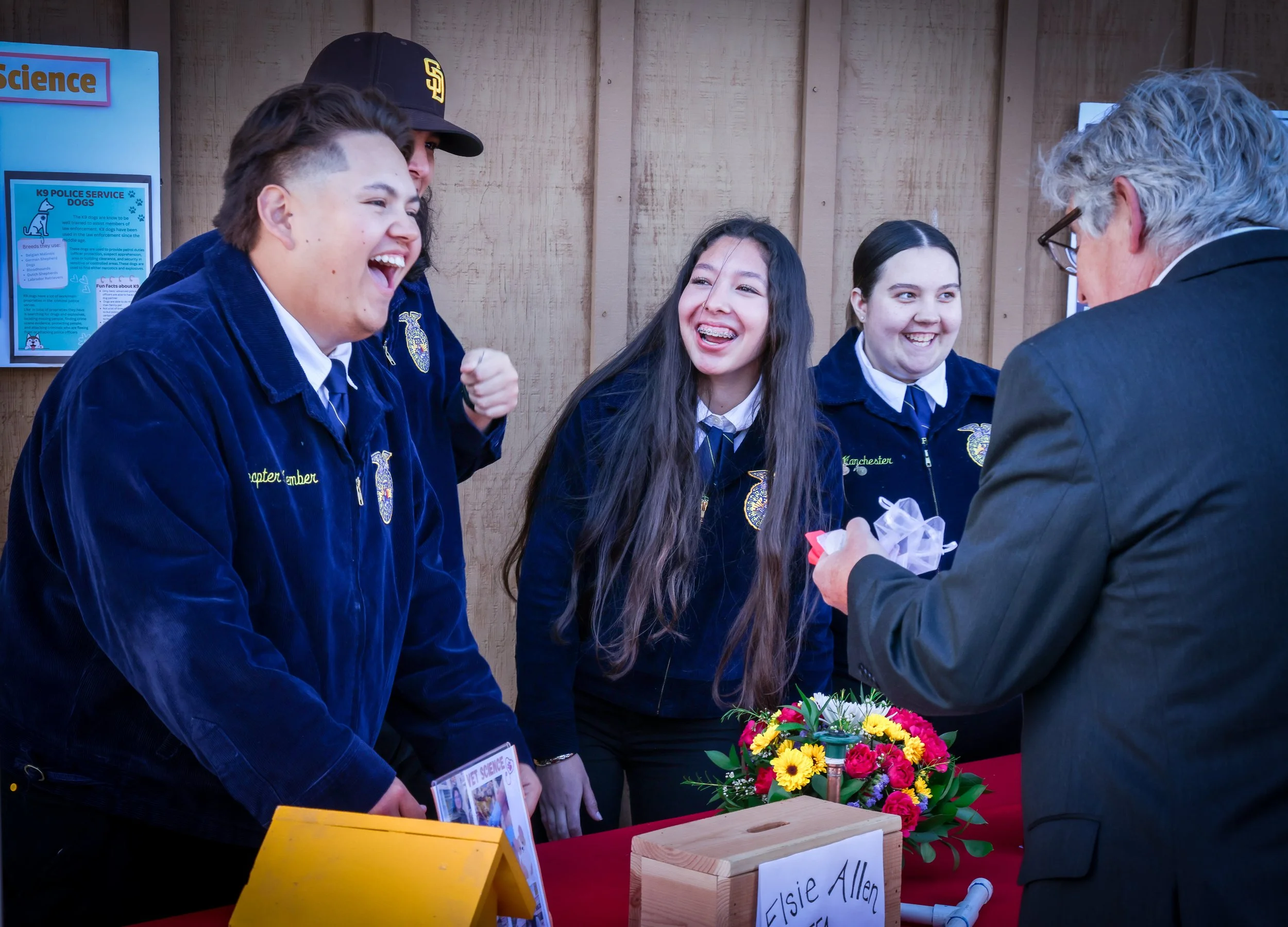 Group of smiling students in blue jackets at a science fair booth, talking to an adult. The booth has a display with flowers and a sign that reads 'Elsie Allen.'