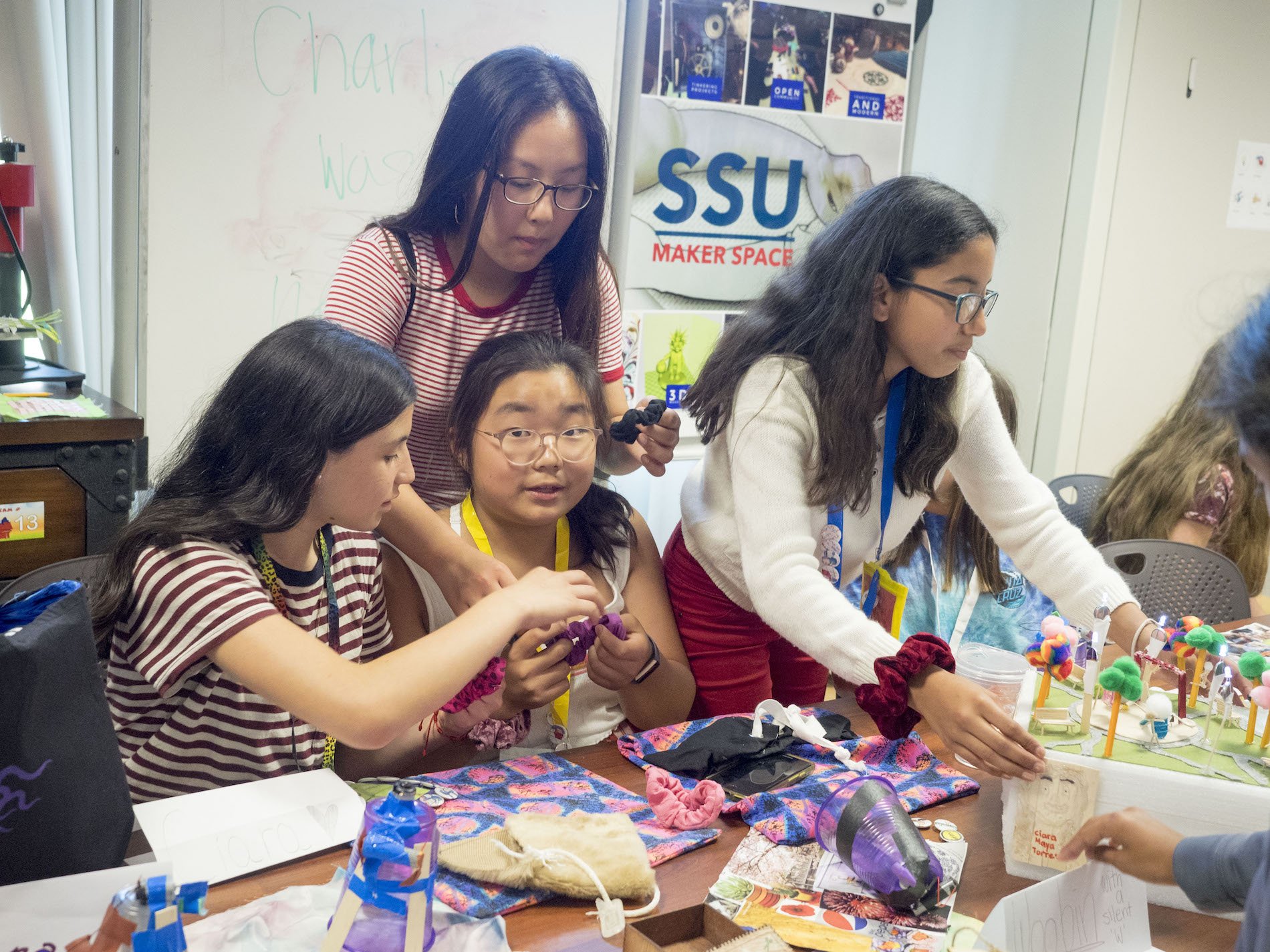 Group of young women working at a craft table in a Maker Space room, with a sign reading 'SSU Maker Space' on the wall behind them. They are creating colorful crafts using various materials.