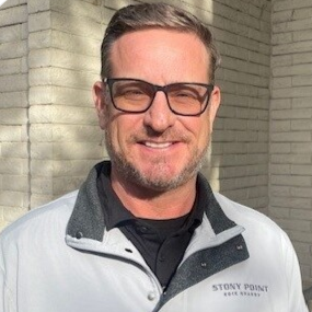 A man with short dark hair, glasses, and facial hair, smiling outdoors against a brick wall, wearing a white and gray jacket.