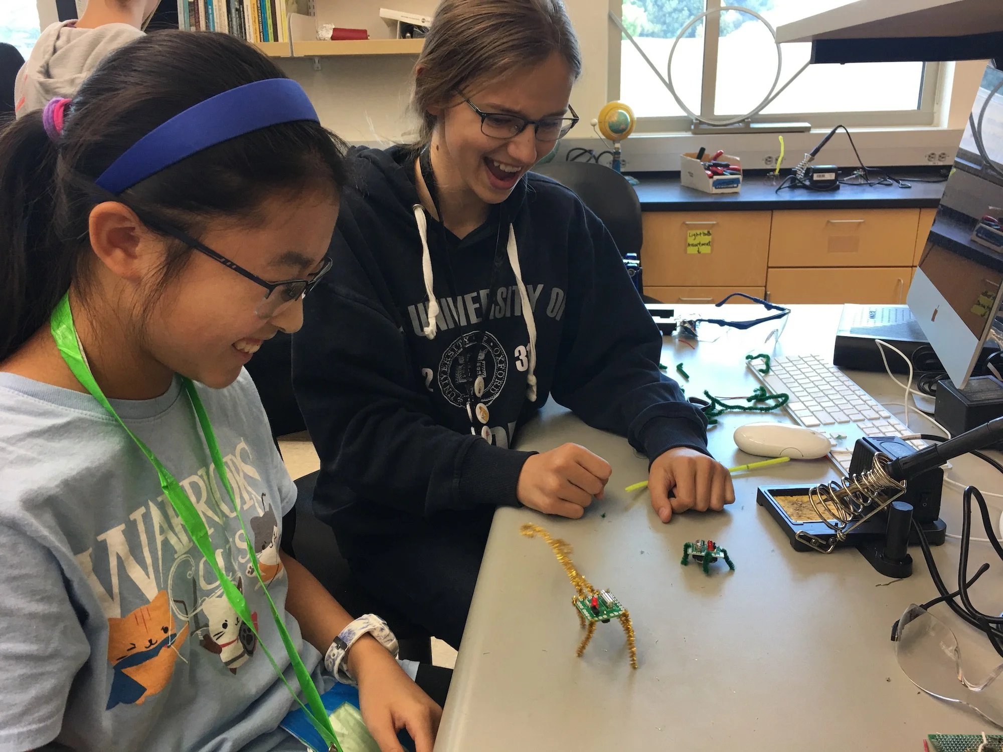 Two students working together at a science or engineering classroom, assembling electronic components or a small project on a green circuit board. They are surrounded by tools, wires, and electronic parts with a computer monitor nearby.