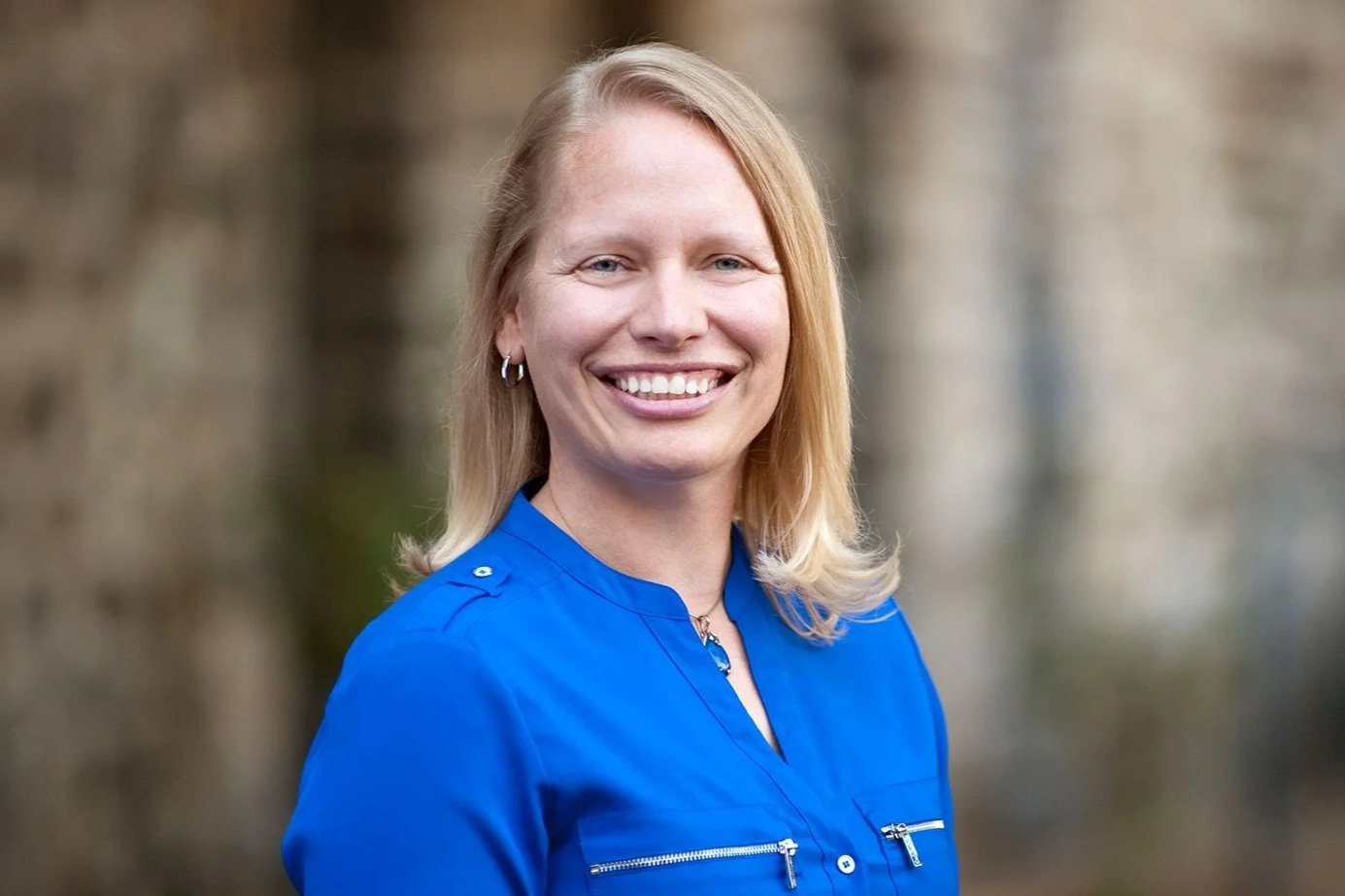 A smiling woman with blonde hair, wearing a bright blue jacket, standing outdoors with a blurred background of trees.