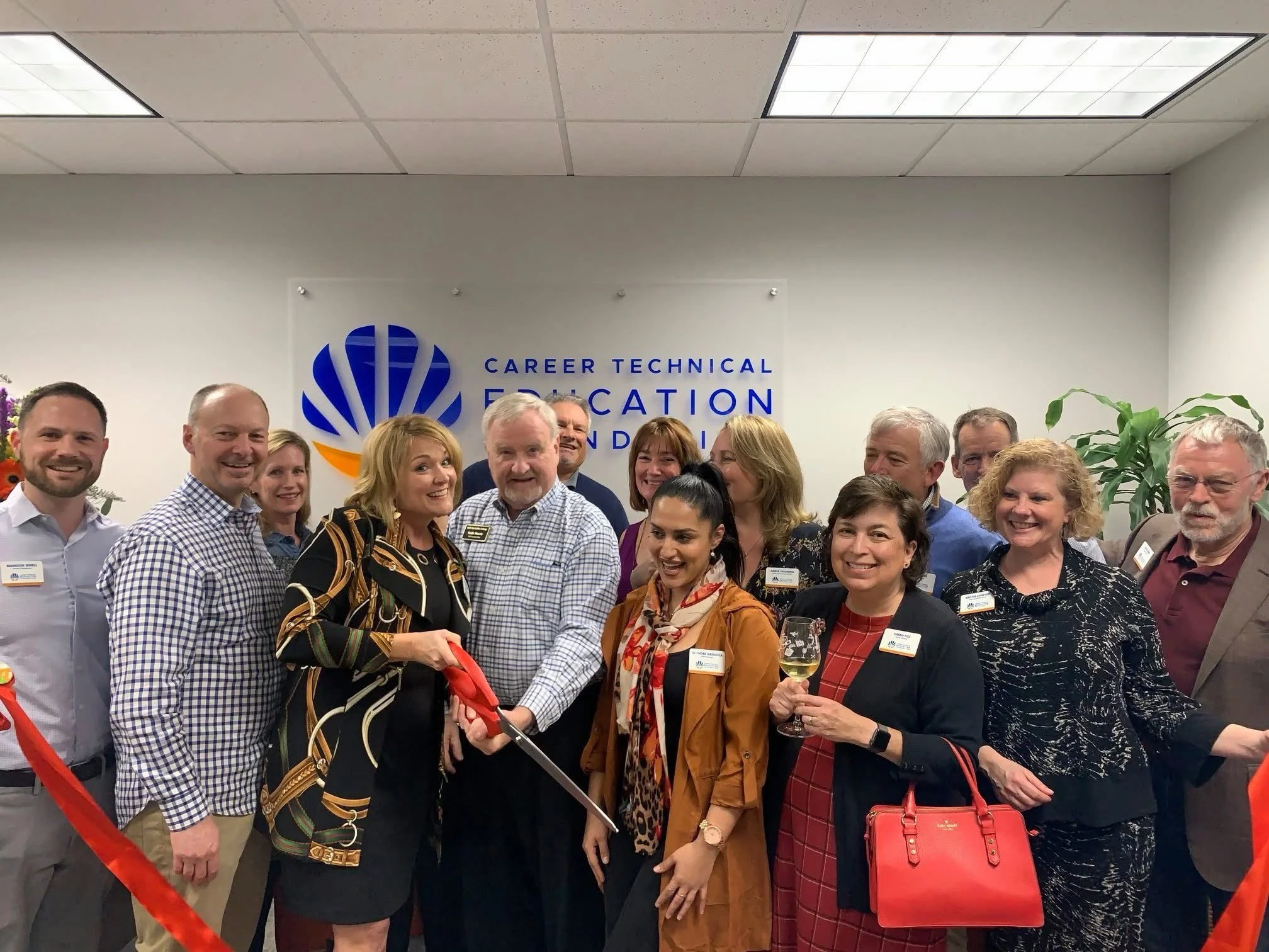 Group of professional men and women gathered at a corporate event, some holding glasses of wine, standing in front of a wall with a sign that reads 'Career Technical Education Foundation' and a blue and orange logo. The group is smiling and posing for the photo.