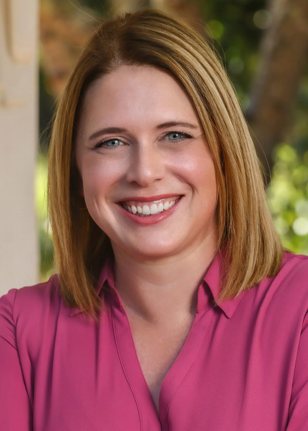 A woman with shoulder-length reddish-brown hair, blue eyes, and a bright smile, wearing a magenta blouse, standing outdoors with greenery in the background.