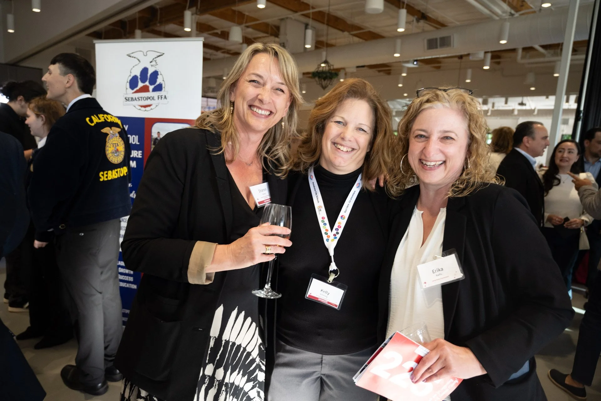Three smiling women with conference name tags at an indoor event, one holding a glass of wine, in front of a banner with a logo and blue, red, and white colors, with other people in the background.
