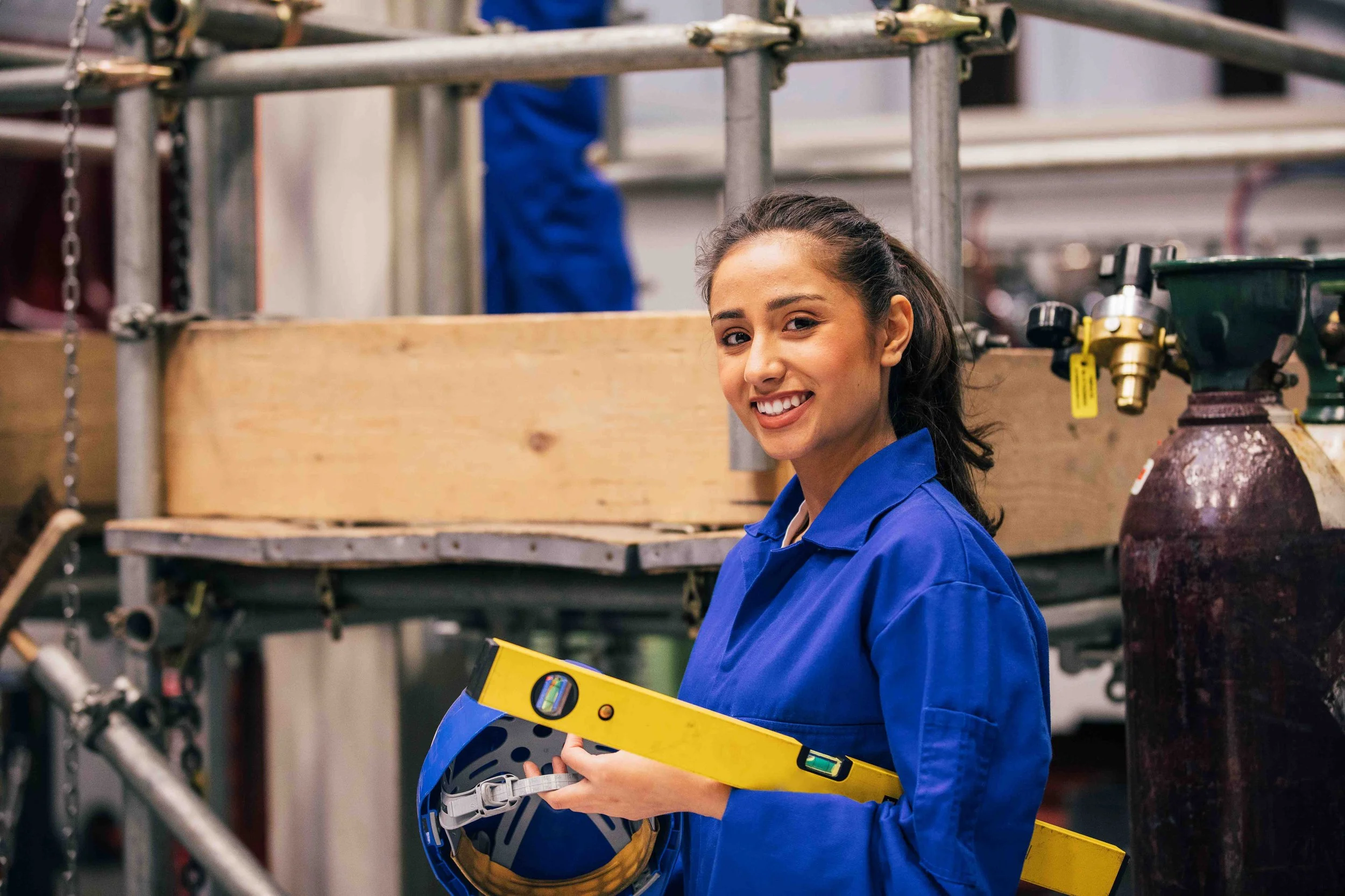 A young woman in blue safety gear smiling in an industrial setting, holding a yellow level and a safety helmet.