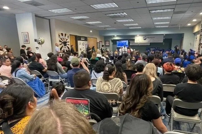 Crowd of people seated and watching a presentation or performance in a large room or conference hall with a stage at the front.