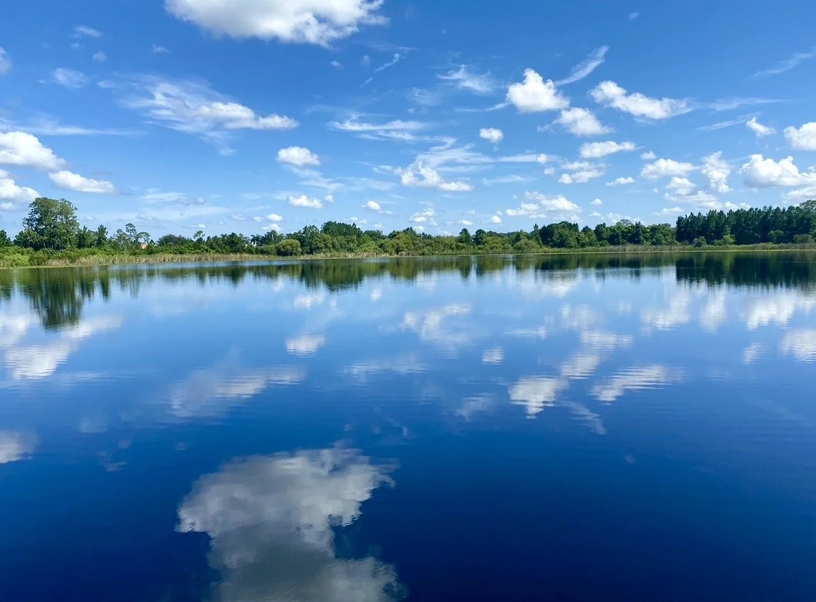 Mirror-like reflection of blue sky and white clouds across a calm lake, photographed by Rob Smith.