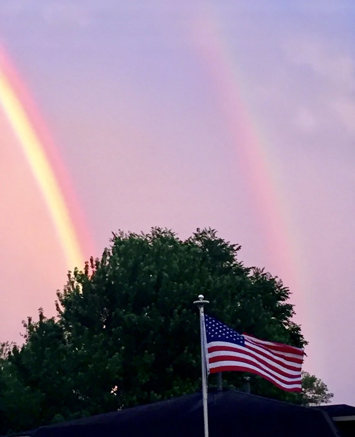 Double rainbow arching above an American flag after a summer storm, captured beneath a colorful evening sky in a photograph by Rob Smith