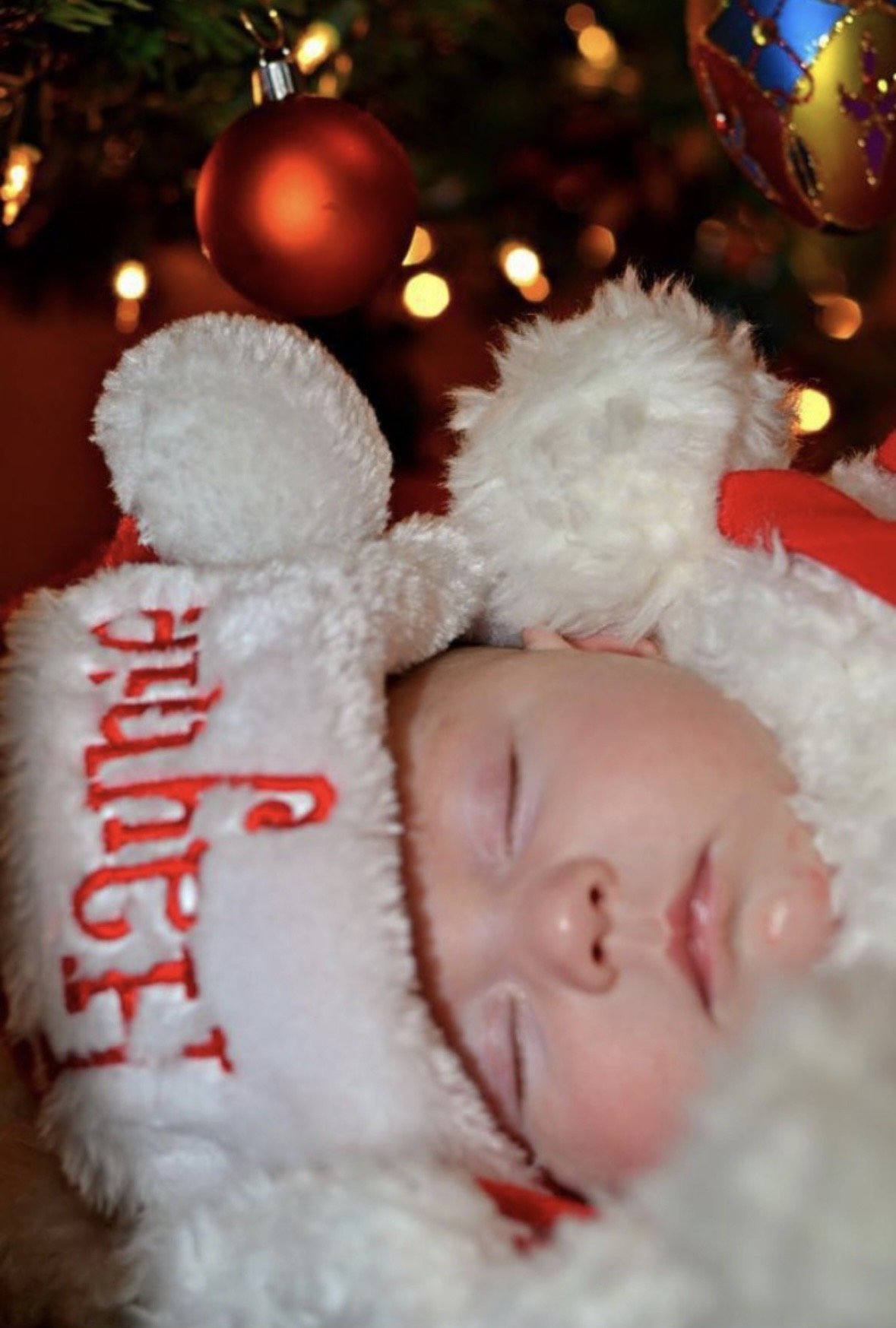 A baby girl dressed in a festive Christmas outfit, peacefully sleeping beneath a decorated Christmas tree in a holiday portrait photographed by Rob Smith.