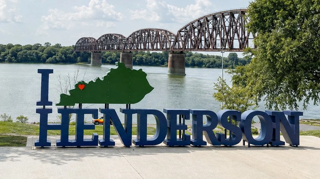 Image of a sign reading Henderson, with an outline of the state of Kentucky and a heart. There is a river and bridge back ground 