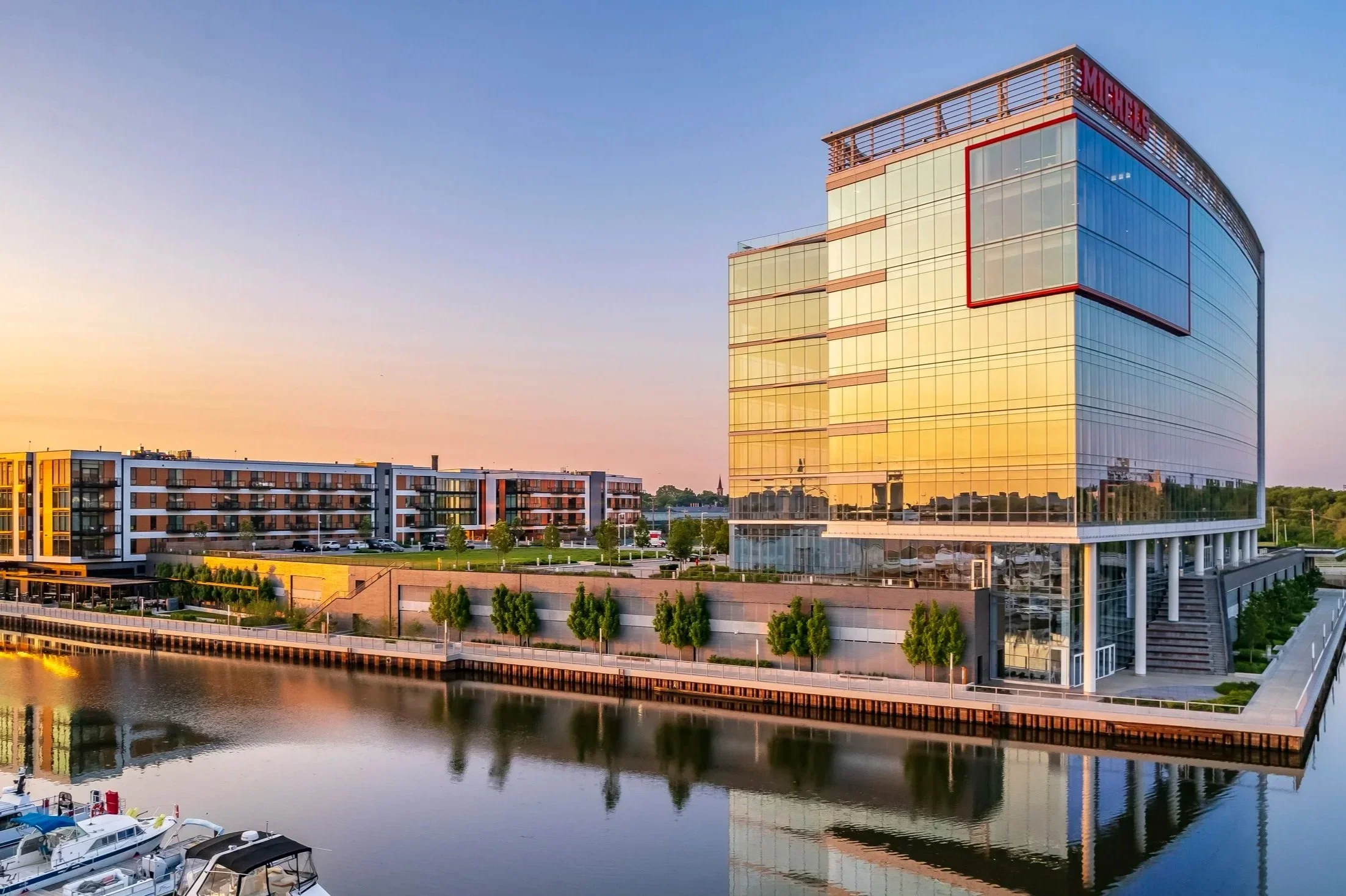 Modern multi-story glass building reflecting the sunset, with residential apartments and boats docked along a river in the foreground.