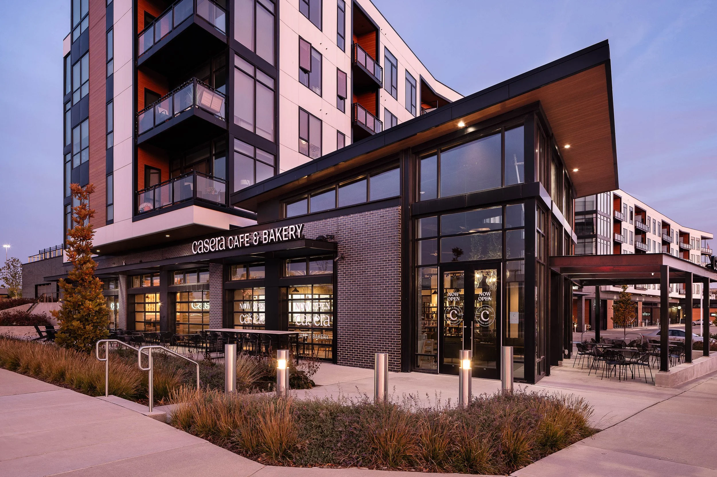A modern multi-story building with a cafe and bakery on the ground floor. The building has large glass windows and balconies on the upper floors. The scene is taken at dusk, with outdoor seating and landscaped area in the foreground.