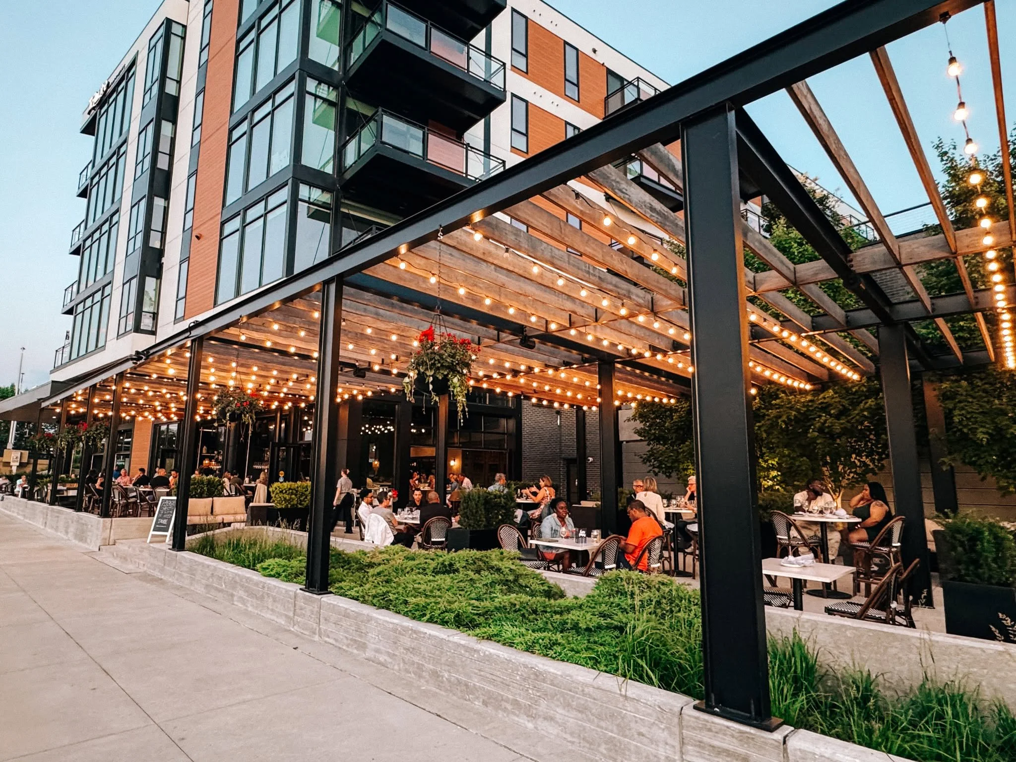 An outdoor dining area of a restaurant or cafe with string lights hanging from a black metal structure. People are seated at tables, dining and socializing, with a modern multi-story building with glass windows in the background.