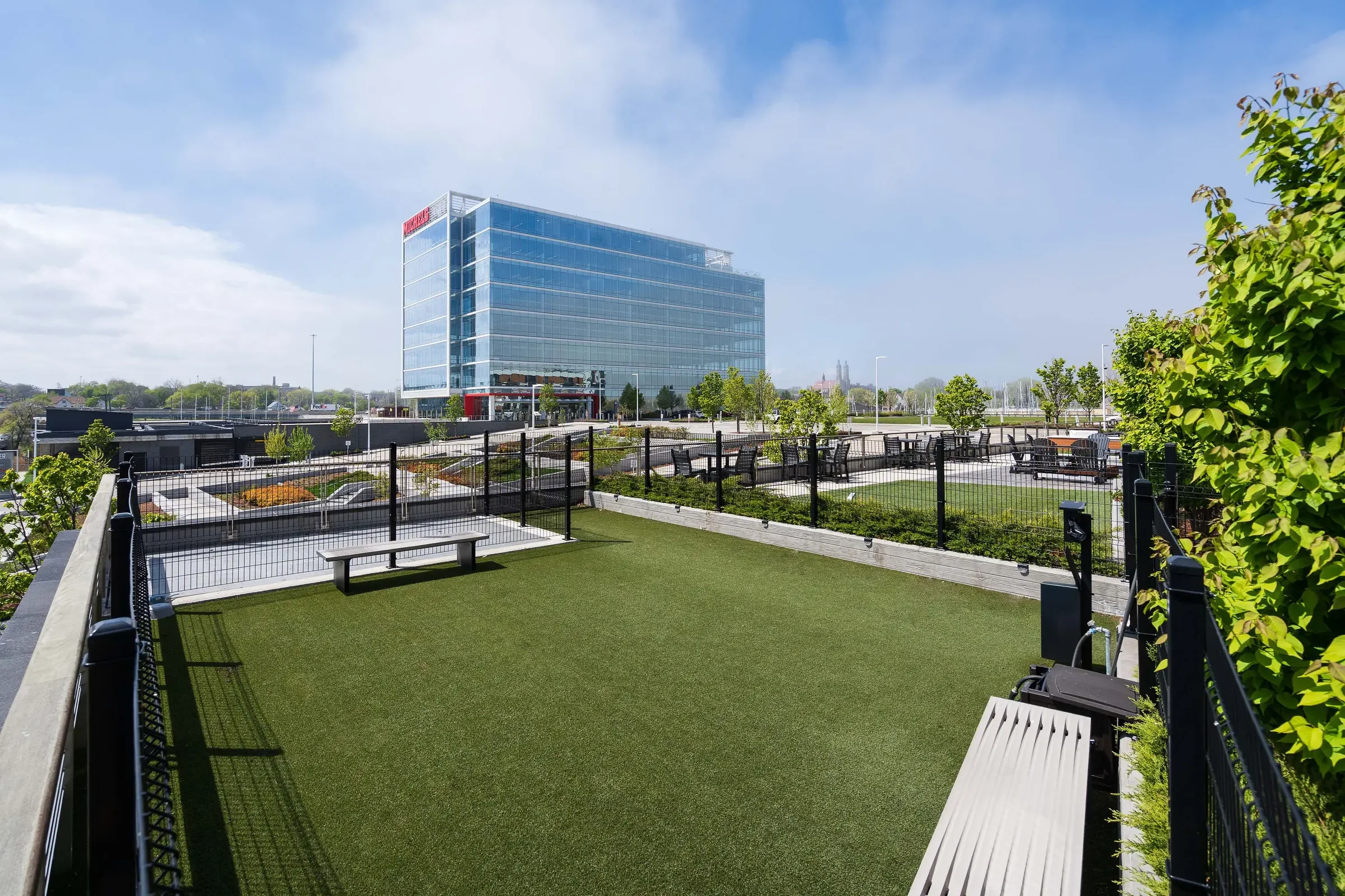 Rooftop garden with benches, small trees, and artificial grass, overlooking modern office building with glass facade and blue sky.