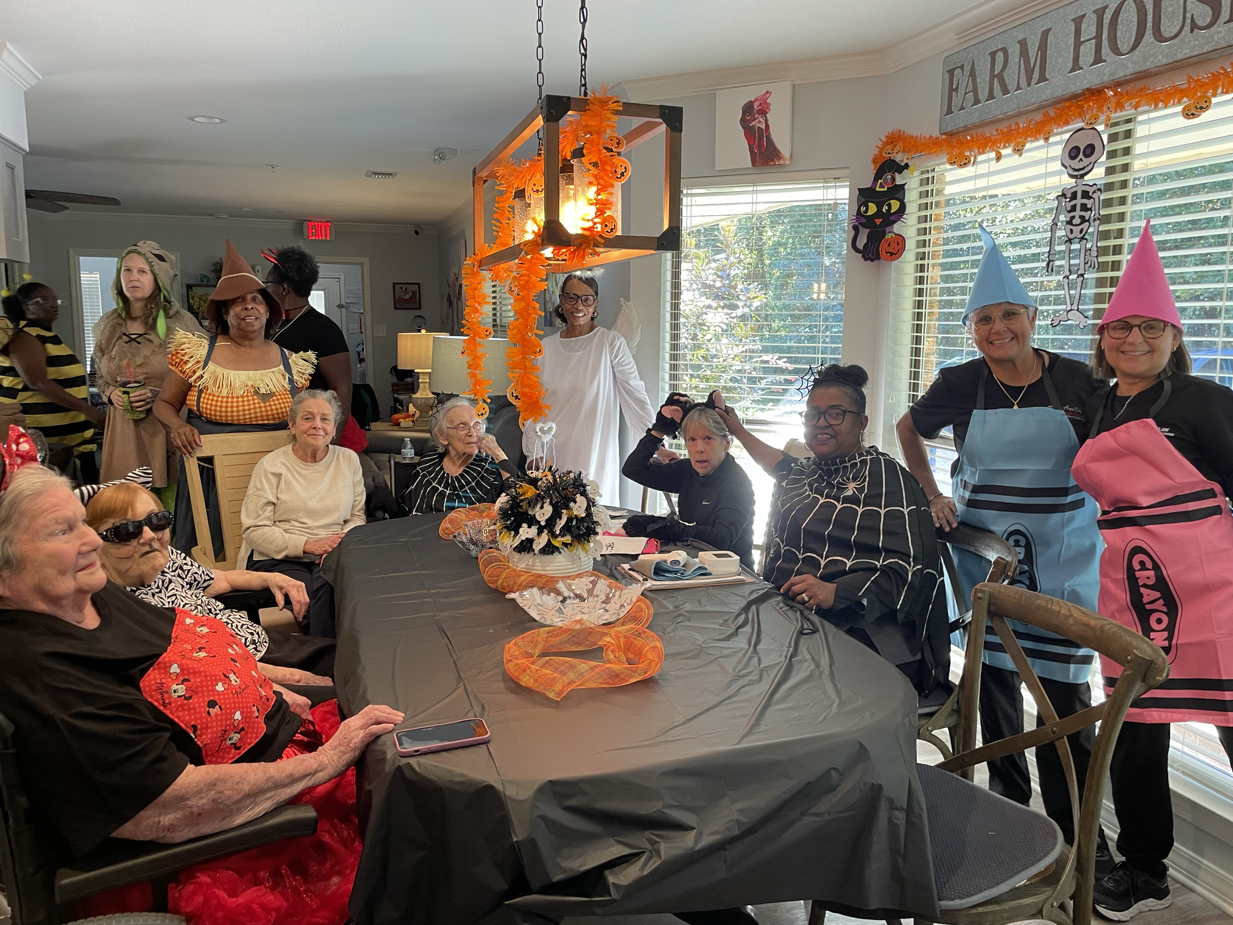 People dressed in Halloween costumes gathered around a decorated table with orange garlands and a Halloween-themed centerpiece, celebrating in a living room with Halloween decorations on the walls.