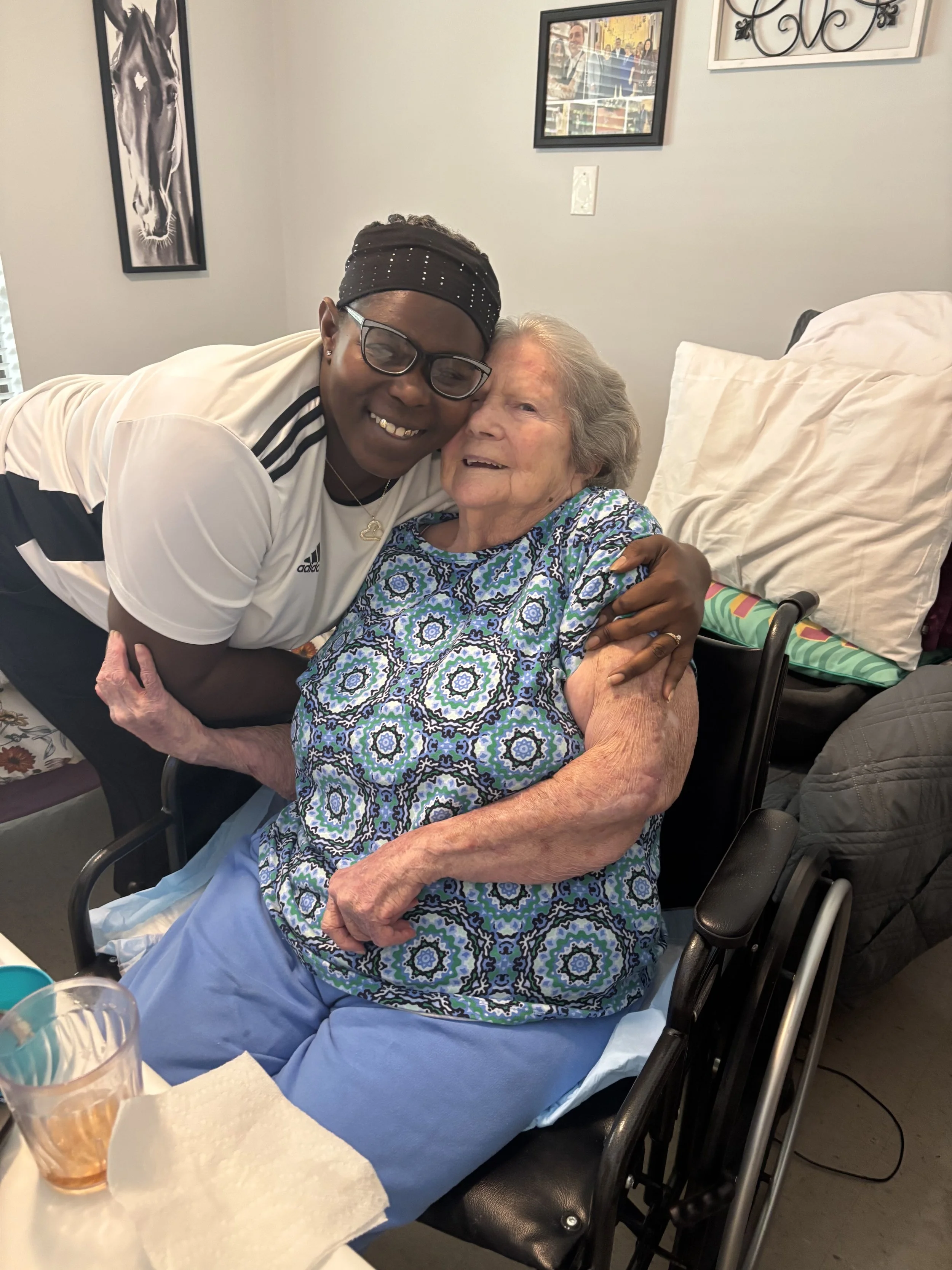 A young woman hugging an elderly woman in a wheelchair, both smiling warmly. The elderly woman is wearing a blue patterned dress, and the room has framed pictures on the wall.