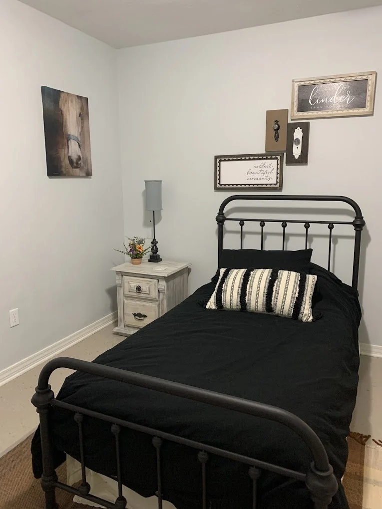 A small bedroom with a black metal bed frame, black bedding, and a rectangular striped pillow. Beside the bed is a white distressed nightstand with a small flower vase, a black lamp, and a gray lamp shade. The wall behind the bed has several decorati