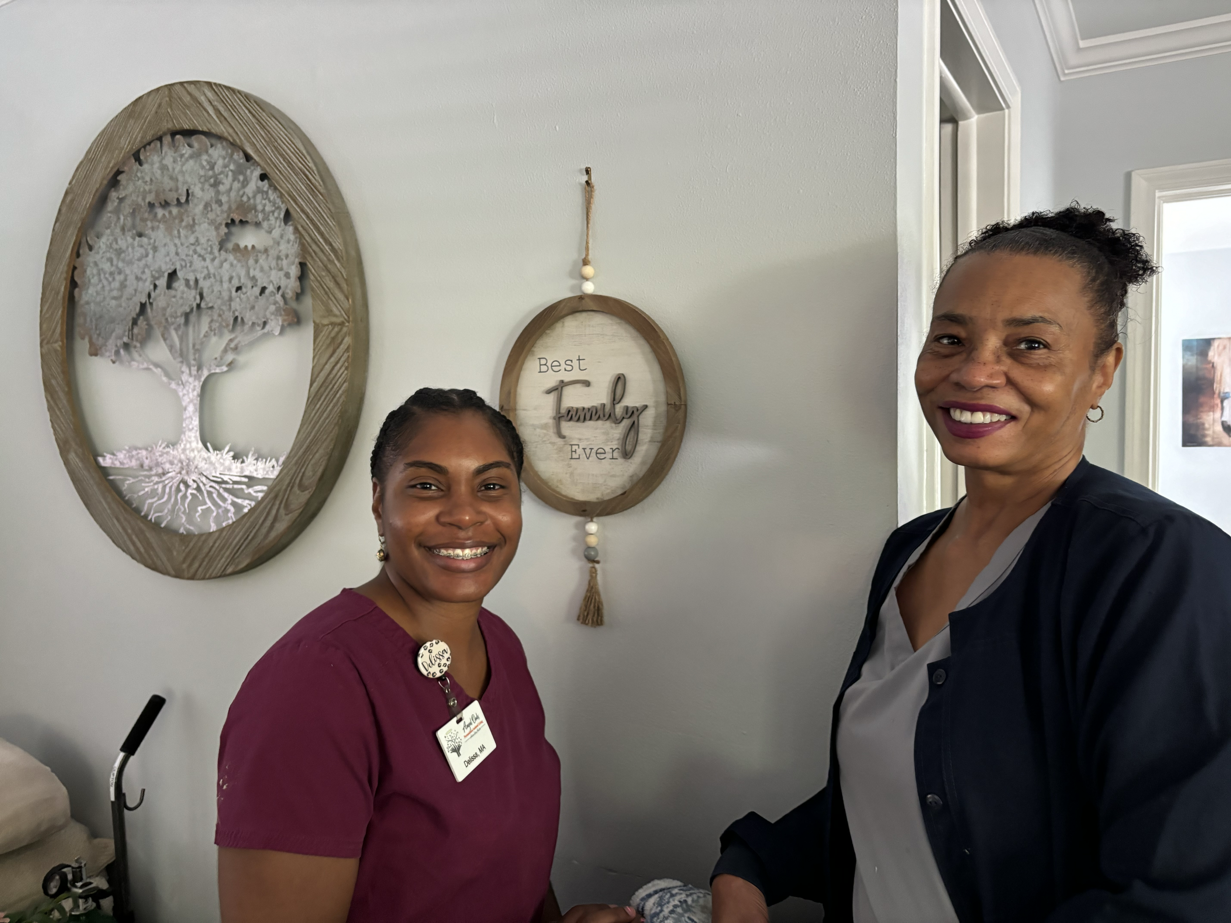 Two women standing next to each other indoors, smiling at the camera. The woman on the left has dark hair and is wearing a burgundy top with a name tag that reads 'Delsey, MIA.' The woman on the right has curly hair pulled back, is wearing a dark jac