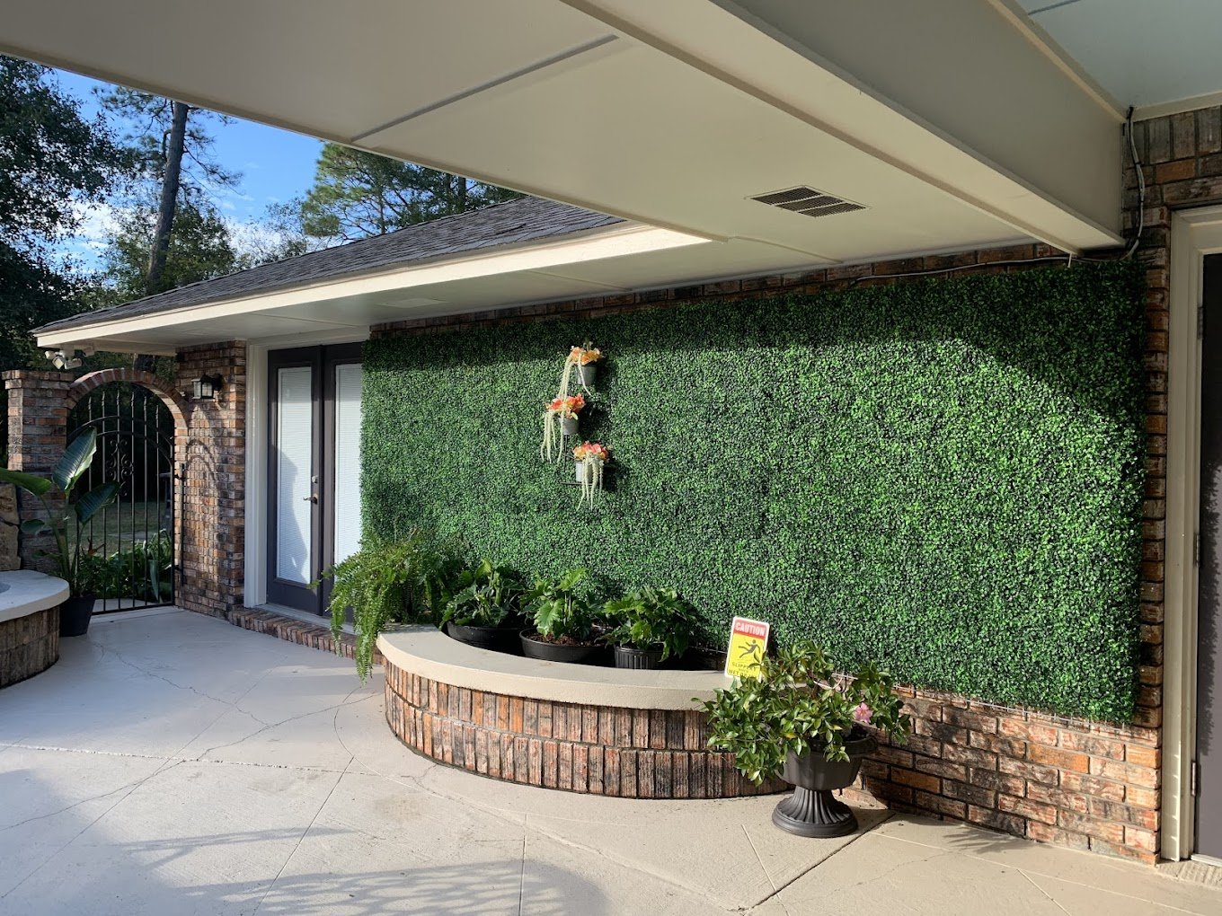 A backyard patio with a green artificial grass wall, potted plants, a brick planter, and decorative flower wall hangings.