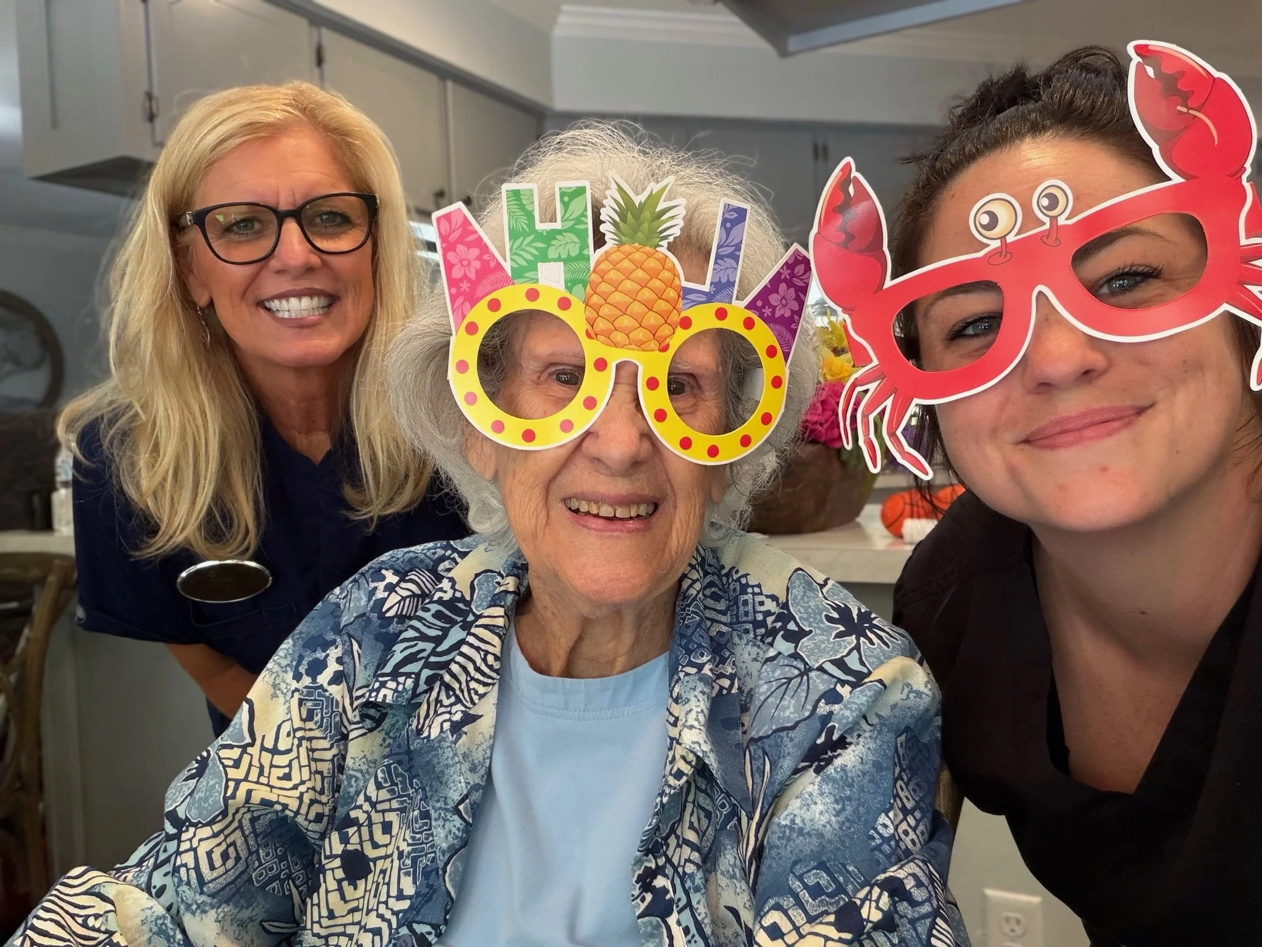 Four women celebrating a fun occasion with playful Hawaiian-themed birthday glasses and a colorful pineapple and floral 'Aloha' sign, smiling at a gathering.