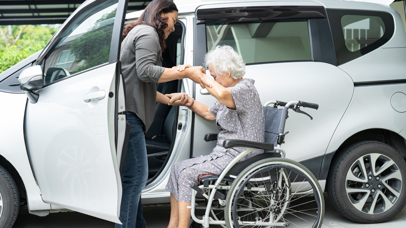 A young woman helping an elderly woman into a wheelchair as she assists her to get into a white car.