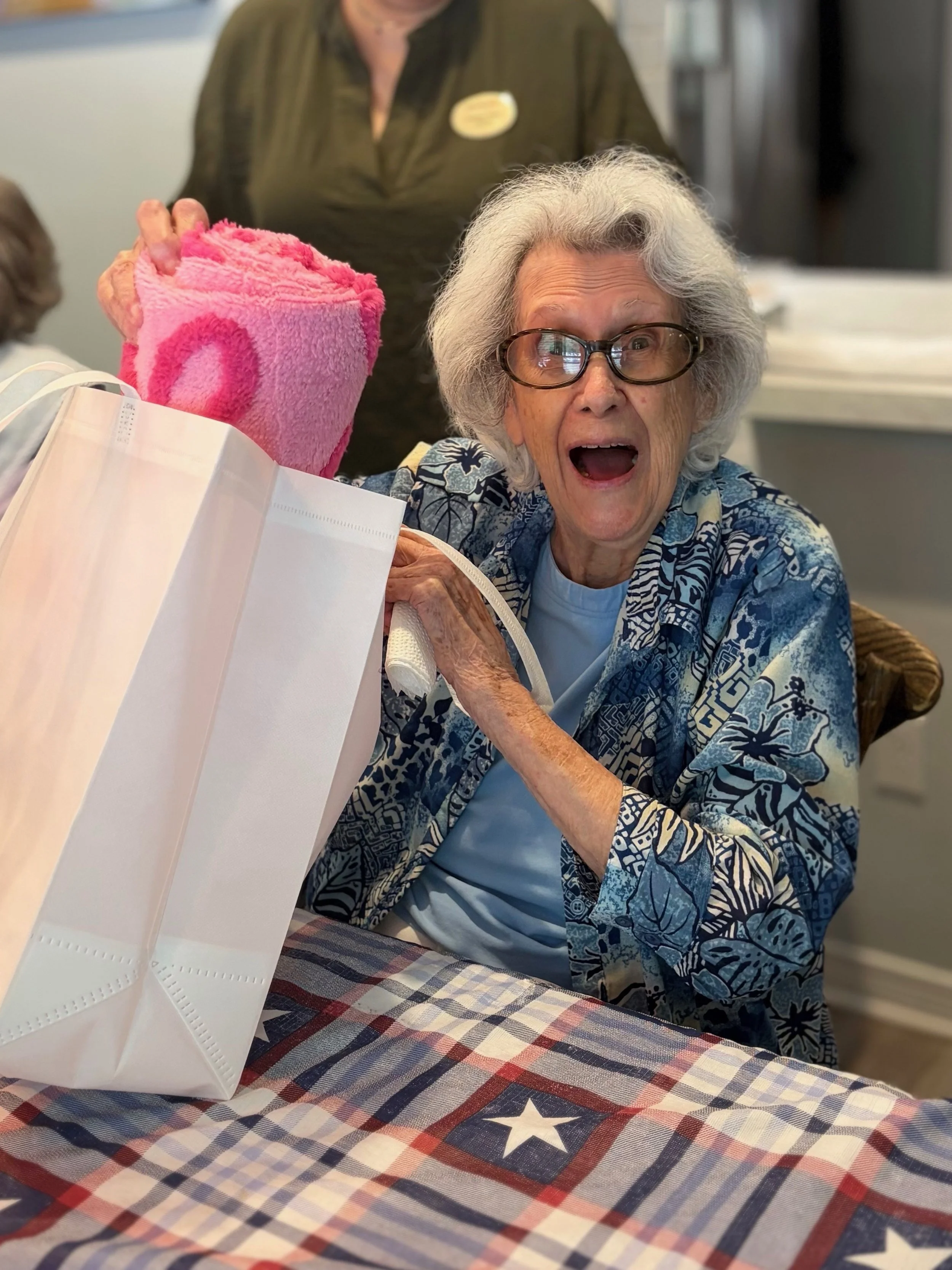 An elderly woman with gray hair, glasses, and an expressive face, sitting at a table with a star-spangled tablecloth, holding a pink blanket, with a nurse or caregiver standing behind her.