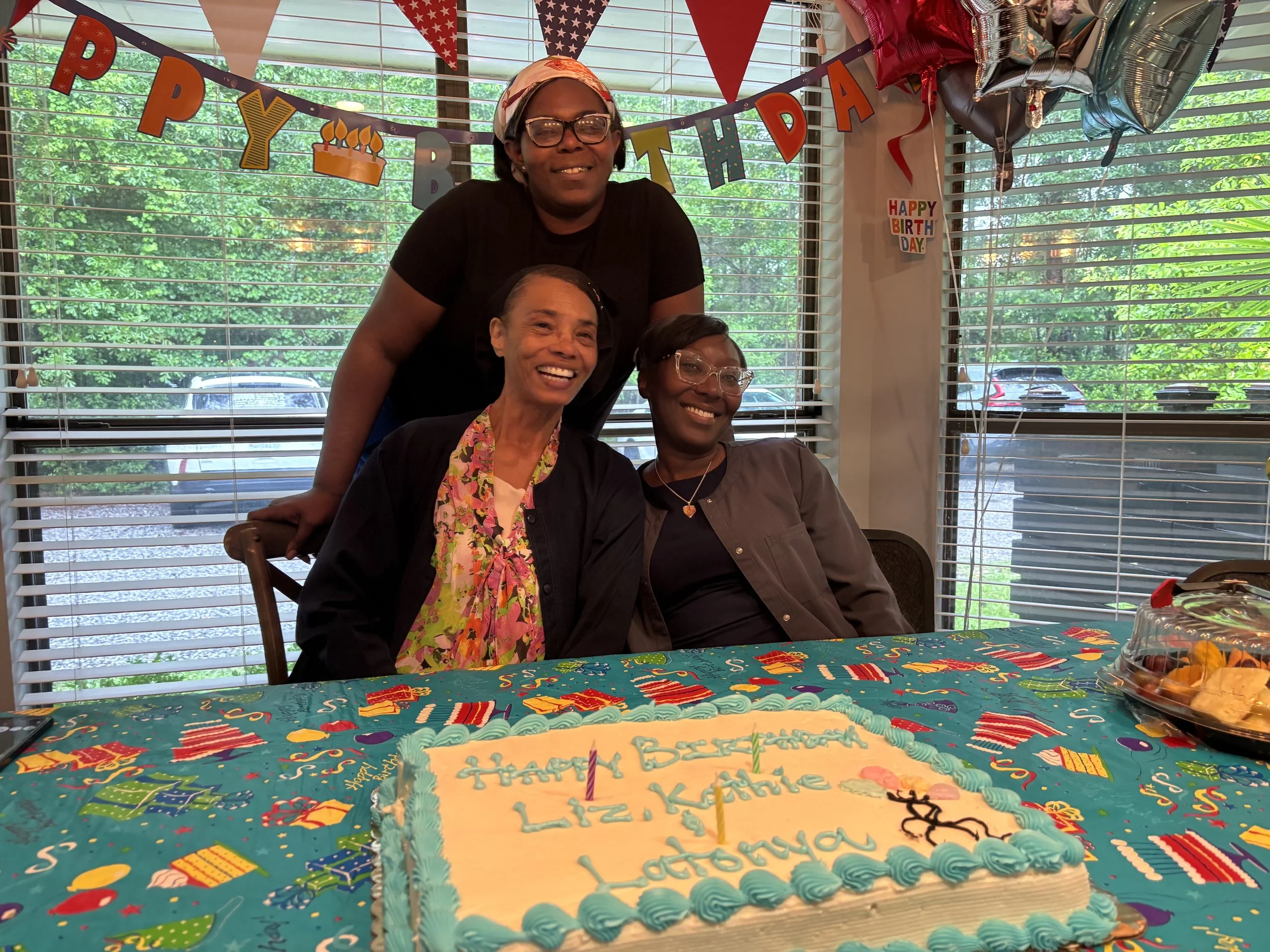 Three women celebrating a birthday at a table decorated with a colorful party tablecloth, a birthday cake with green frosting and candles, and a platter of snacks. They are smiling with a window and decorated banners in the background that say 'Happy