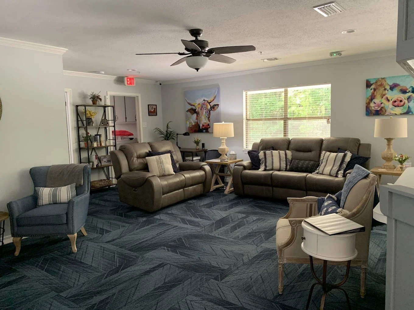 Living room with beige leather sofas, a blue armchair, patterned pillows, a ceiling fan, two table lamps, wall art including paintings of a cow and pigs, a window with blinds, a black bookshelf with decorative items, and a blue patterned carpet.