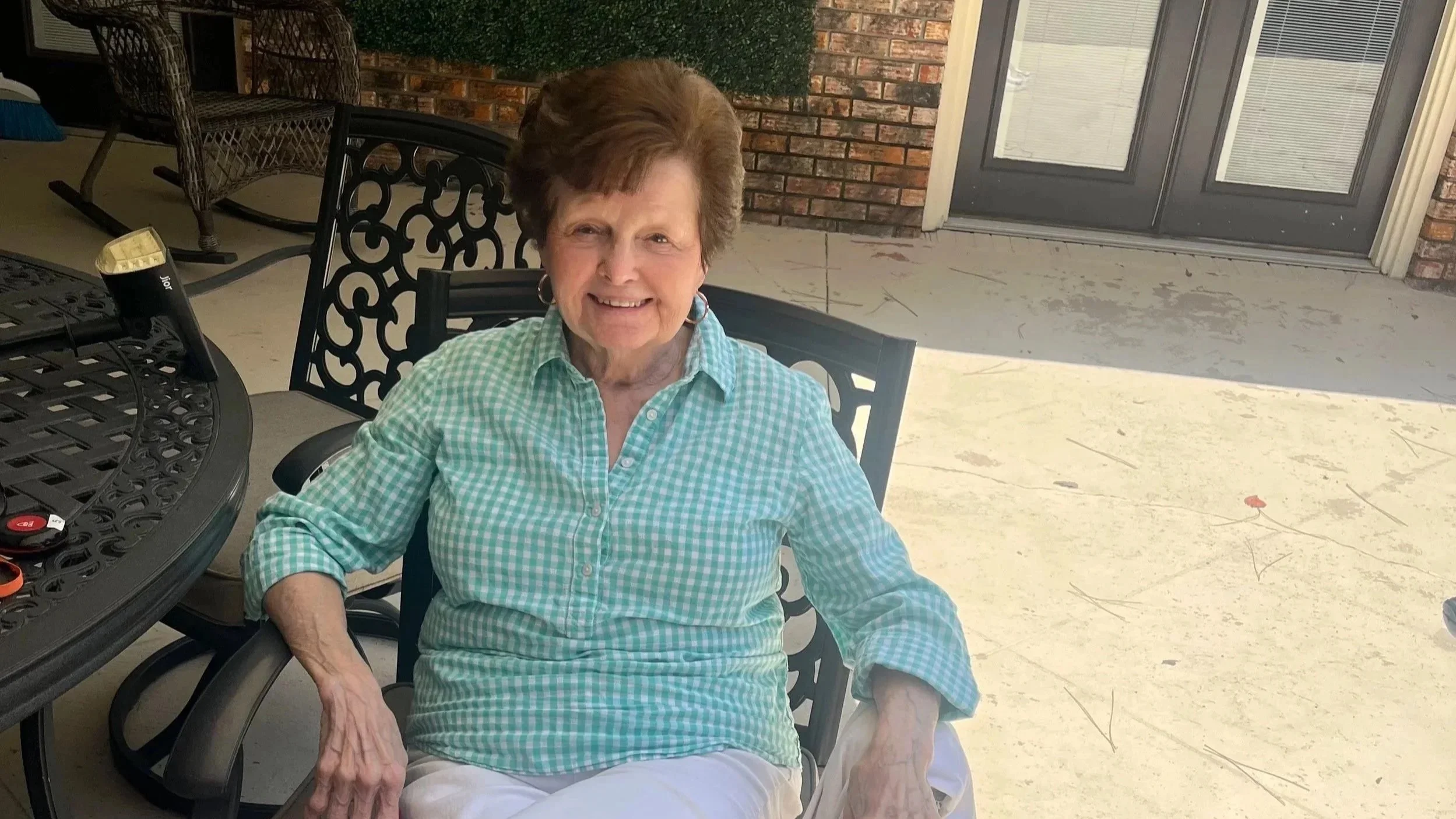 An elderly woman with short brown hair, wearing a green and white checkered shirt, seated outdoors at a black wrought iron table with matching chairs, smiling at the camera. Angel Oaks has a beautiful courtyard area to relax and unwind.