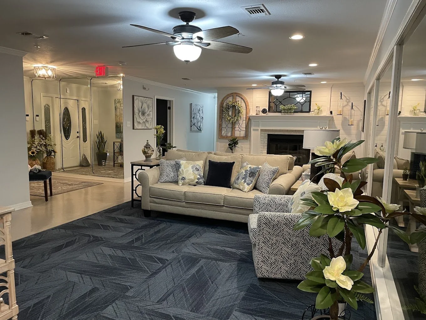 A cozy living room with a beige sofa decorated with patterned pillows, a fireplace with shelving and decorative items, ceiling fans, and plants. A glass wall with reflections separates the living space from an entryway with double doors and a mirror.