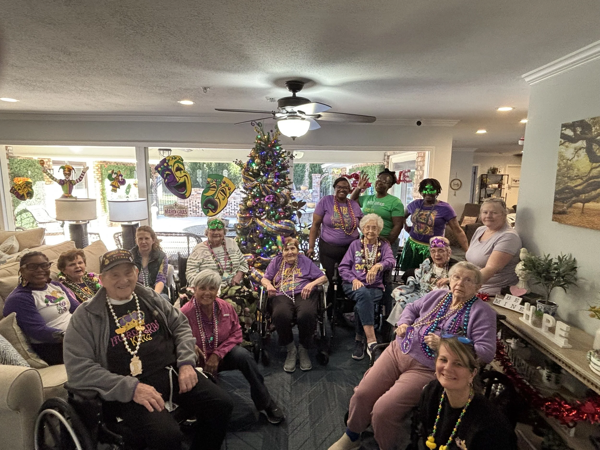 Group of elderly women and caregivers celebrating Mardi Gras in a living room decorated with Mardi Gras masks, a Christmas tree with colorful lights, beads, and a festive atmosphere.