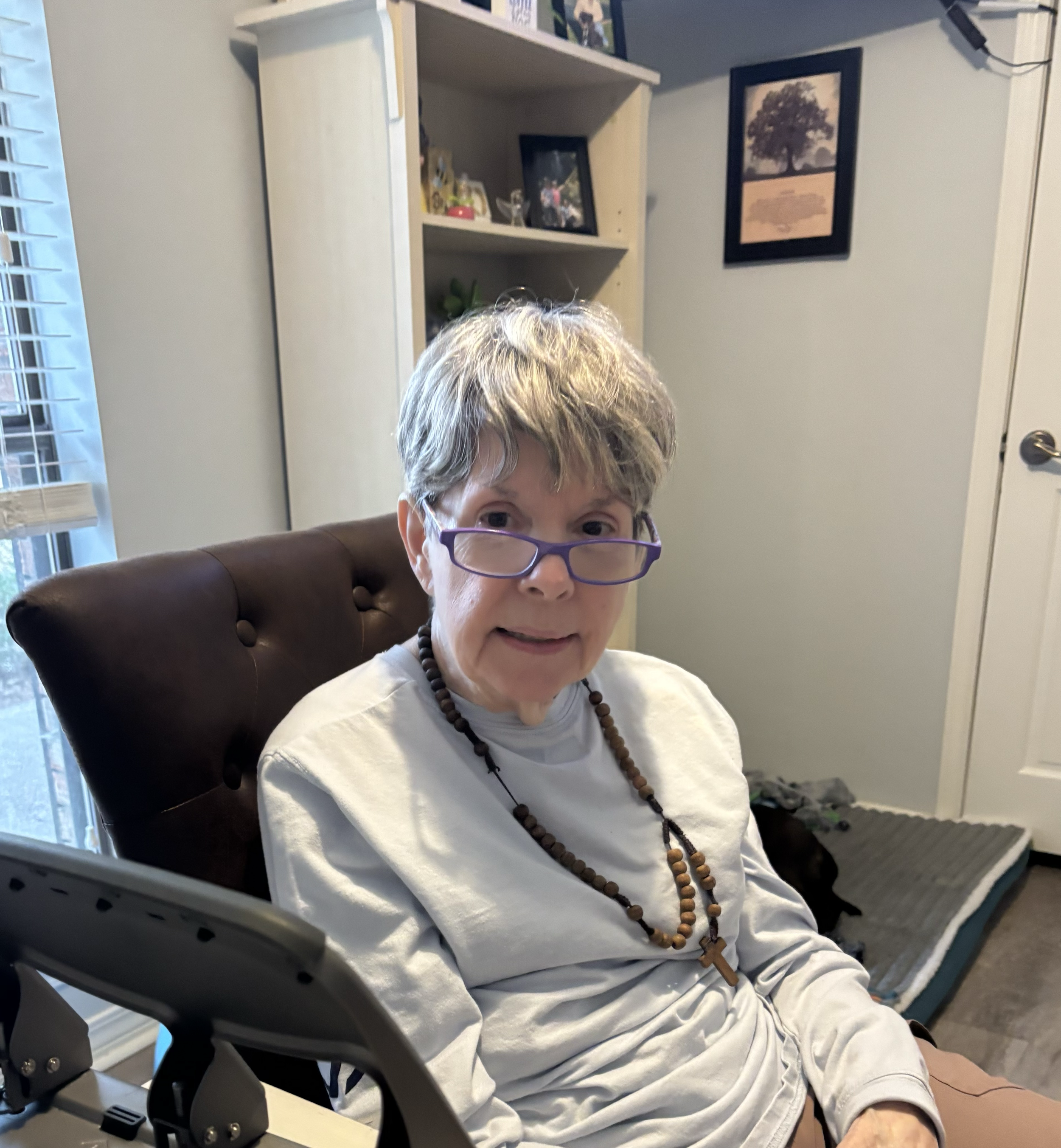 A woman with gray hair and purple glasses sitting in a brown leather chair, wearing a white shirt and a wood bead necklace with a cross pendant, in a room with a shelving unit and framed pictures on the wall.