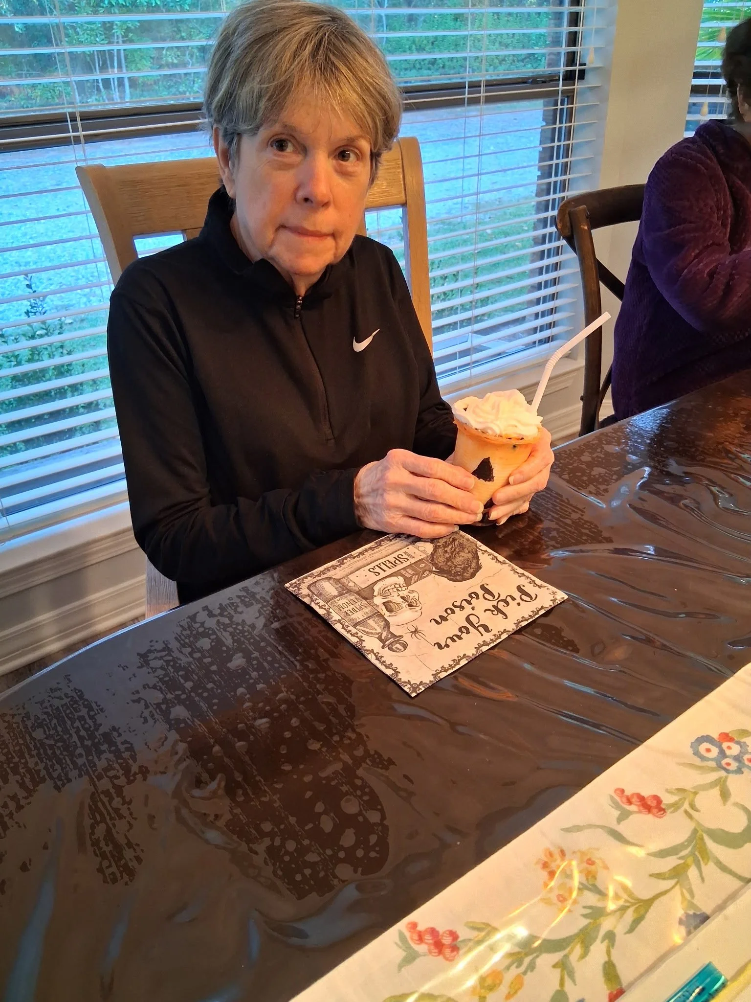 An elderly woman with short gray hair sitting at a wooden table, holding a bowl of ice cream with whipped cream and a straw. She wears a black Nike sports jacket. Behind her are window blinds and a view of green plants outside.