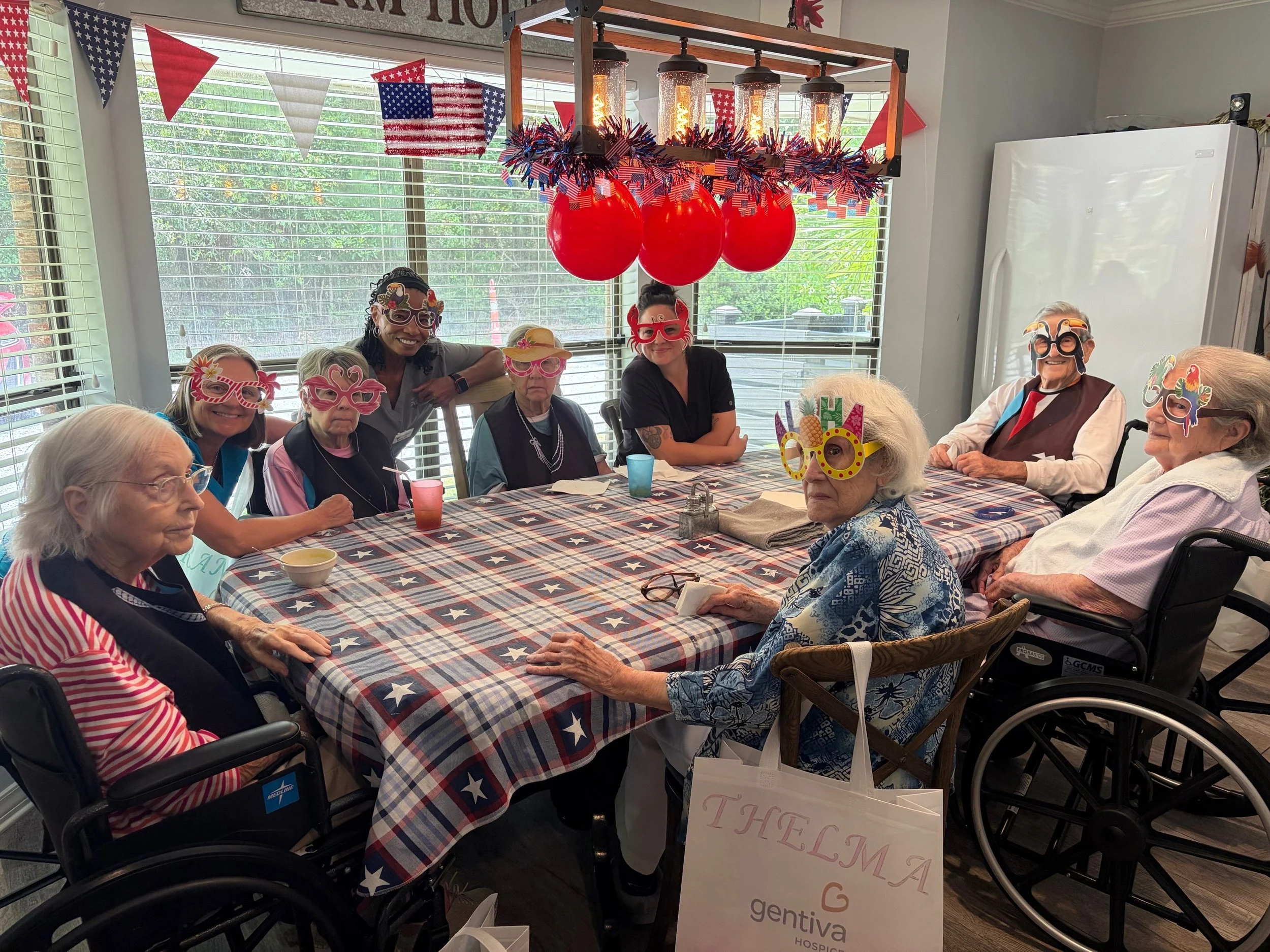 A group of elderly and middle-aged people wearing holiday-themed masks celebrating around a table decorated with an American flag tablecloth. The background has patriotic bunting, red balloons, and a hanging light fixture with exposed bulbs.