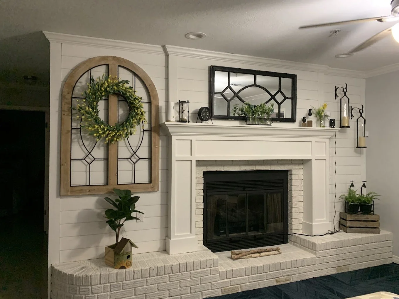 Living room fireplace with a white brick hearth, decorated with a wreath on a window frame, potted plants, candles, and decorative items on the mantle, with a large mirror above.