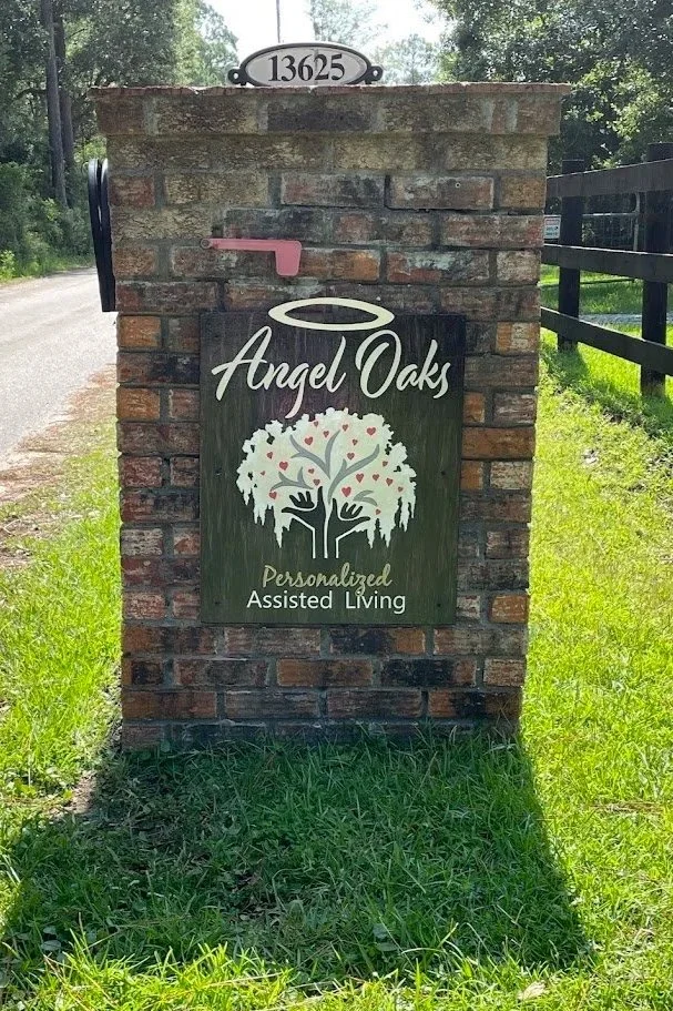 A brick sign with the address 13625 on top. It advertises 'Angel Oaks,' a personalized assisted living facility, with a logo of a tree with a halo and hands underneath.