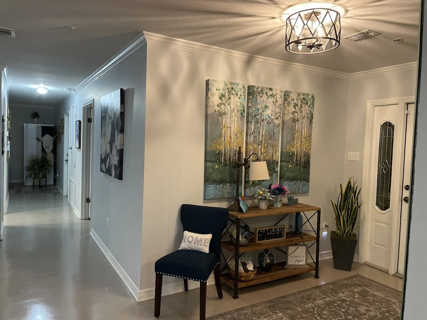 Interior view of a home entryway with a front door, a wooden console table decorated with flowers and a sign, a navy blue chair with a pillow, a large potted plant, and hanging artwork of a forest scene. The hallway leads to other rooms with addition