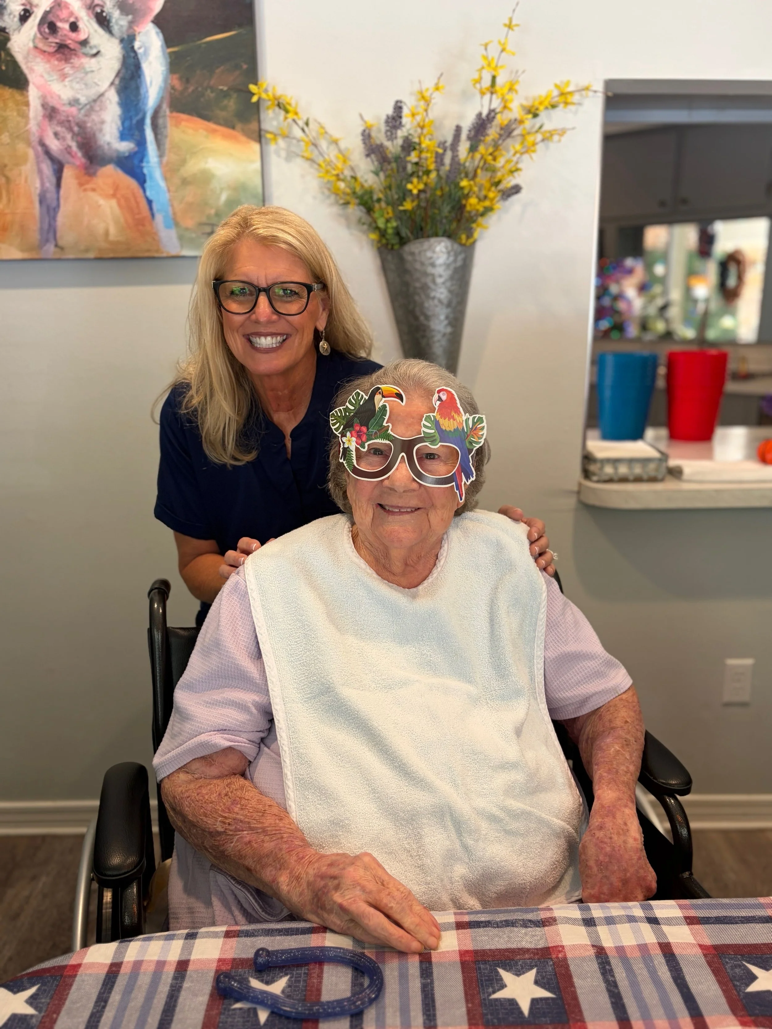 A woman in a wheelchair wearing novelty glasses with colorful toucans and tropical leaves, smiling, while another woman stands behind her, smiling, in a room decorated with a large pig painting, yellow flowers in a silver vase, and a table with a che
