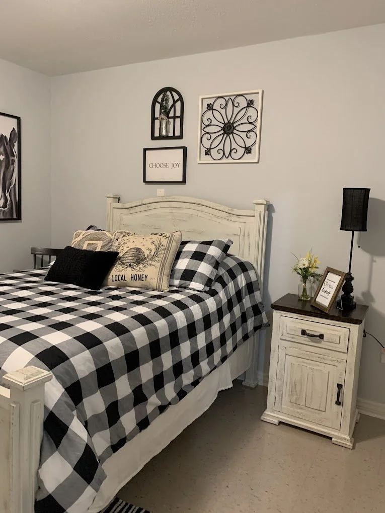 A cozy bedroom with a white wooden bed frame and black-and-white checkered bedding. Decorative pillows are on the bed, including one with a honey theme and a black pillow. To the right, a white nightstand holds a small flower arrangement, a framed si