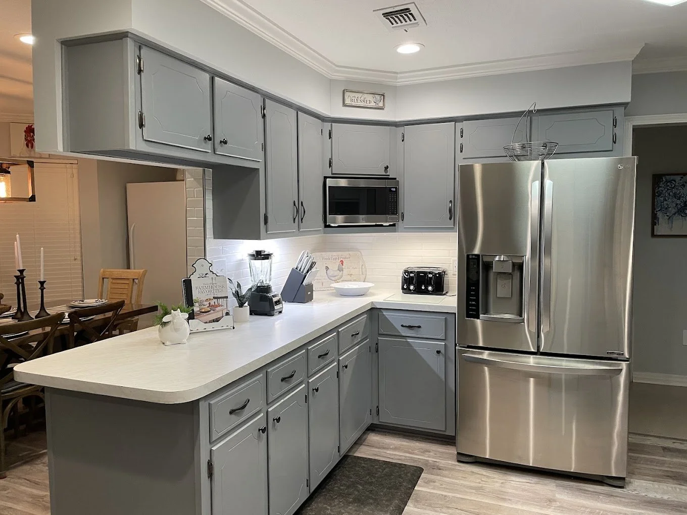 A kitchen with gray cabinets, a white countertop, and stainless steel appliances including a refrigerator, microwave, and toaster. There is a small dining area with wooden chairs and a table on the left side.