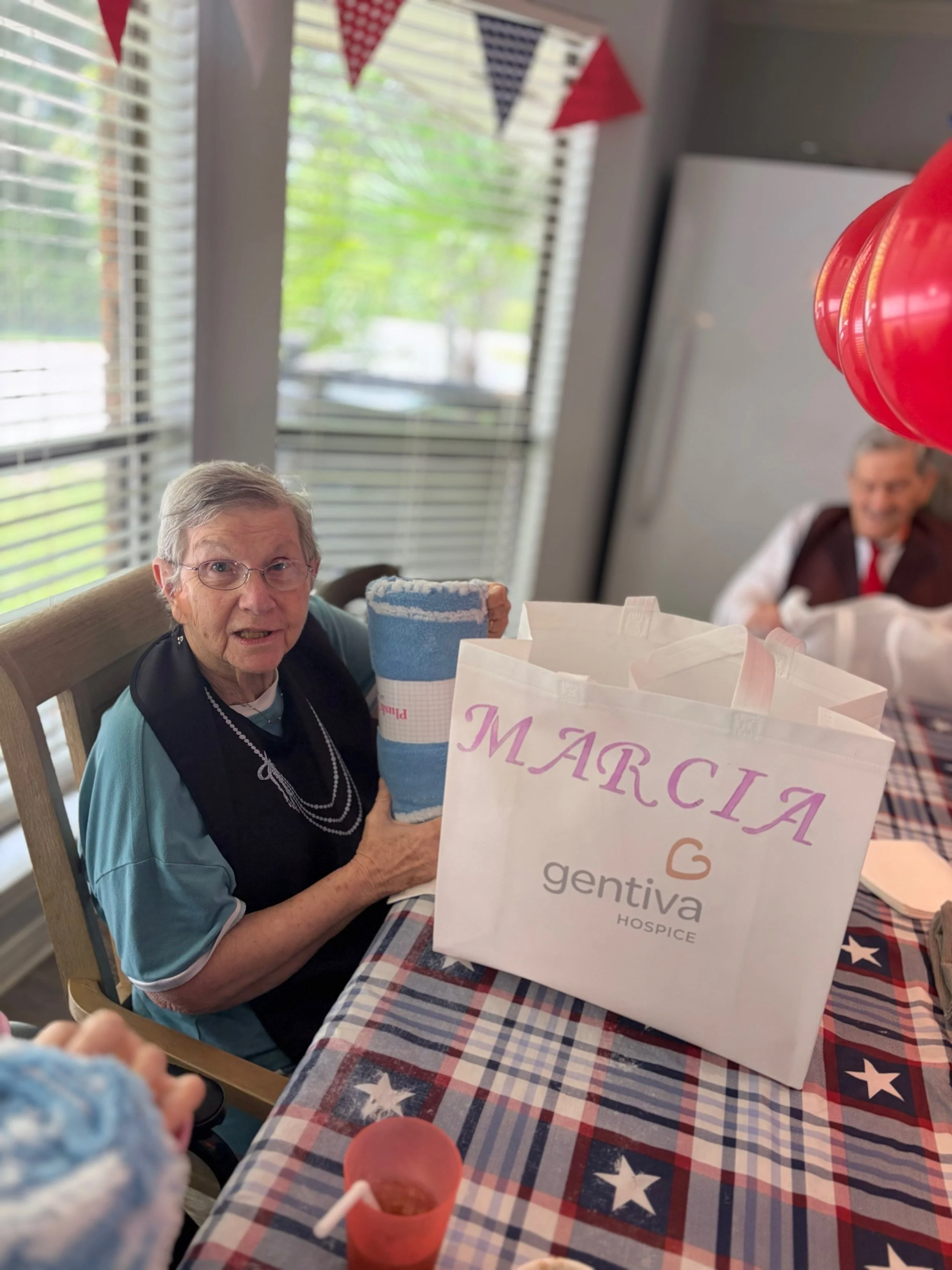 An elderly woman with glasses sitting at a table with a tablecloth featuring stars and stripes, holding a blue towel. On the table is a white gift bag with the words 'MARCIA' and 'gentiva hospice,' a red cup with a drink, and other items. In the back