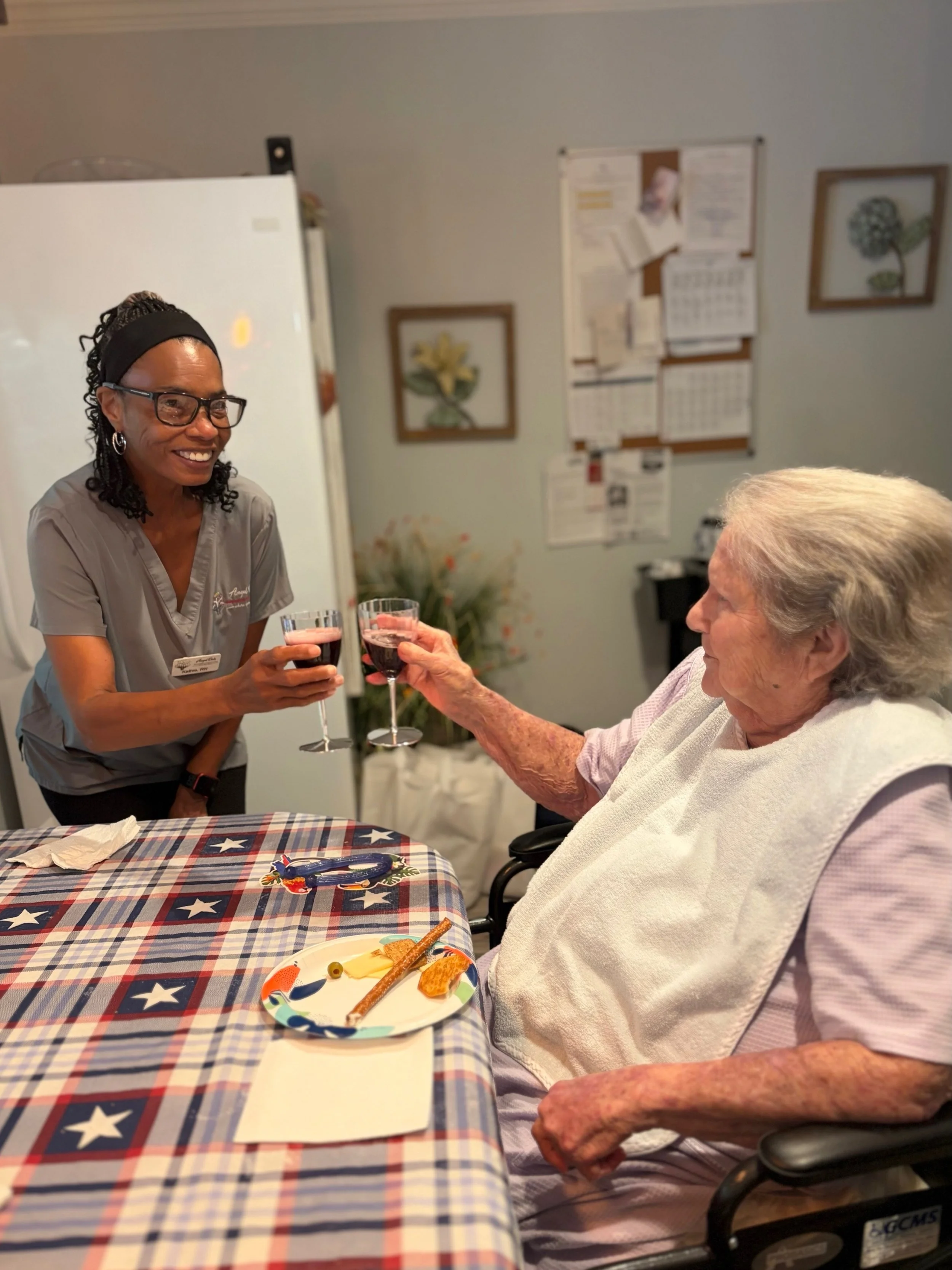 A young woman in scrubs and glasses is smiling and handing a glass of red wine to an elderly woman in a wheelchair, who is wearing a light-colored shirt and a white blanket, in a room with patriotic-themed tablecloths, framed artwork of flowers, and 