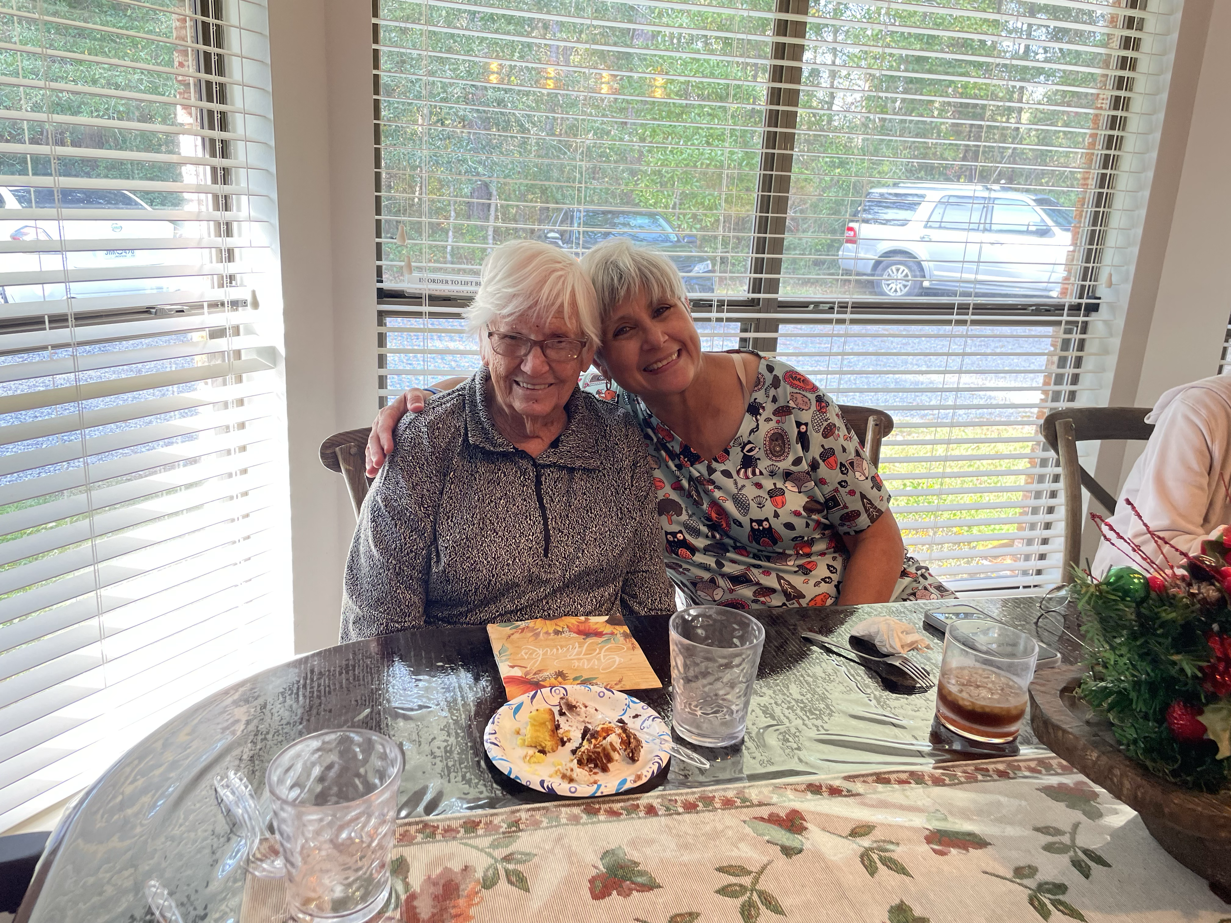 Two smiling women sitting at a table during a celebration or meal, with food, drinks, and a floral centerpiece on the table. Large windows with white blinds reveal parked cars and greenery outside.