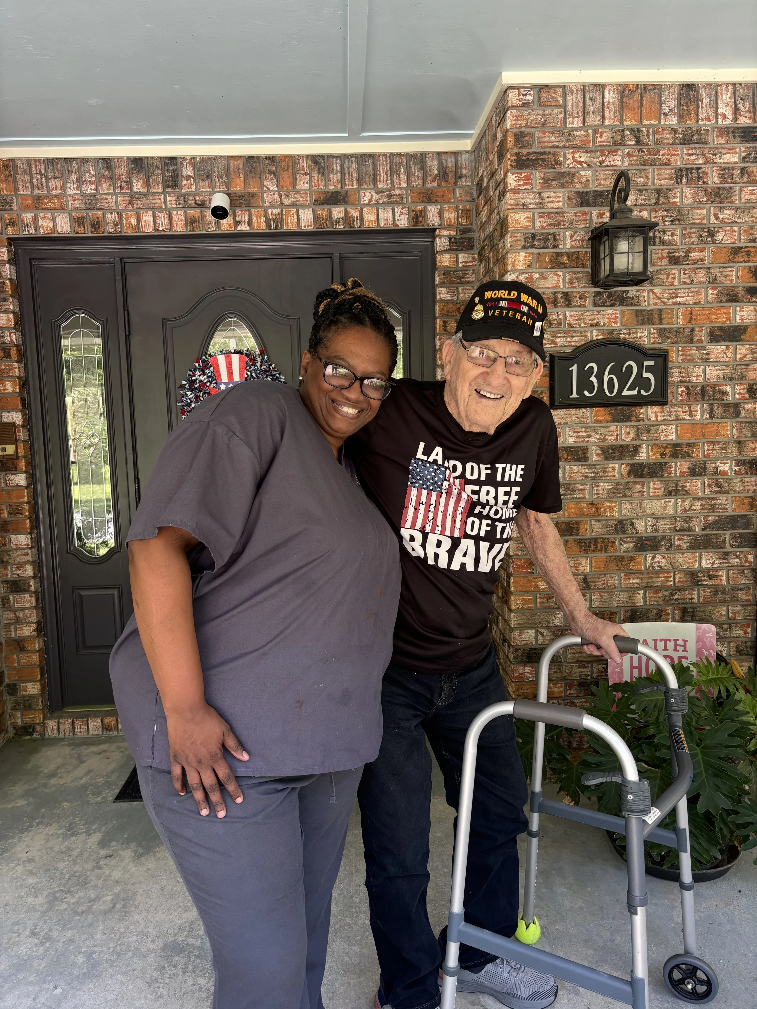 A woman in gray medical scrubs stands beside an elderly man wearing a black 'World War II Veteran' hat, a black t-shirt with patriotic writing, and glasses. The man is using a walker, and they are standing outside a house with a brick wall and a blac