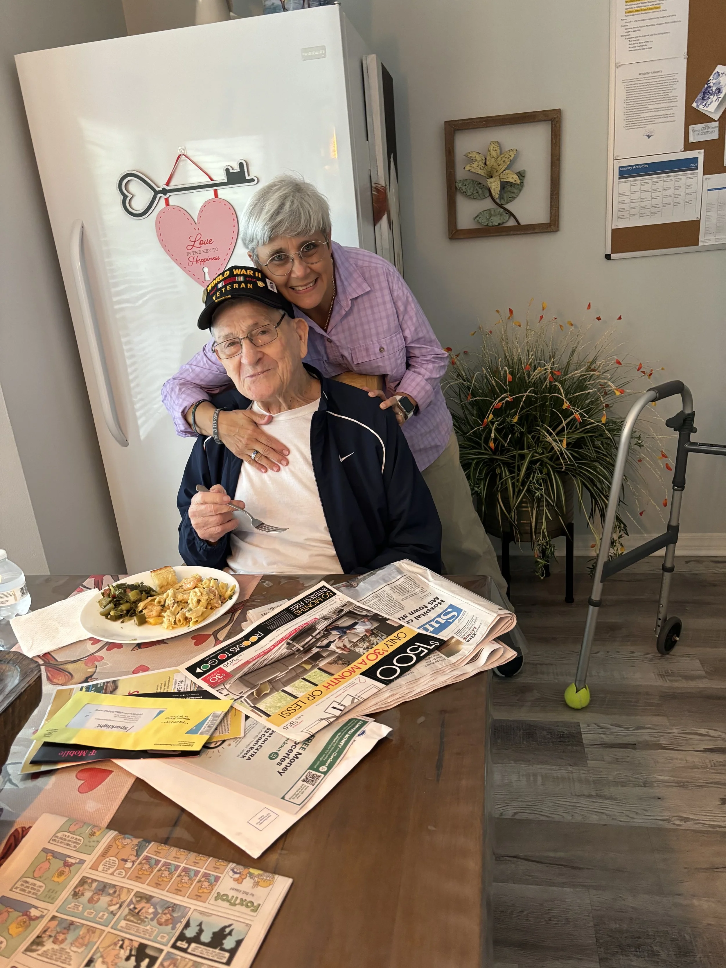 A woman hugging an elderly man wearing a World War II veteran cap, seated at a table with newspapers, comic strips, and a plate of food, in a room with a refrigerator decorated with a heart and key magnet, a wall with framed leaf art, and a large pla